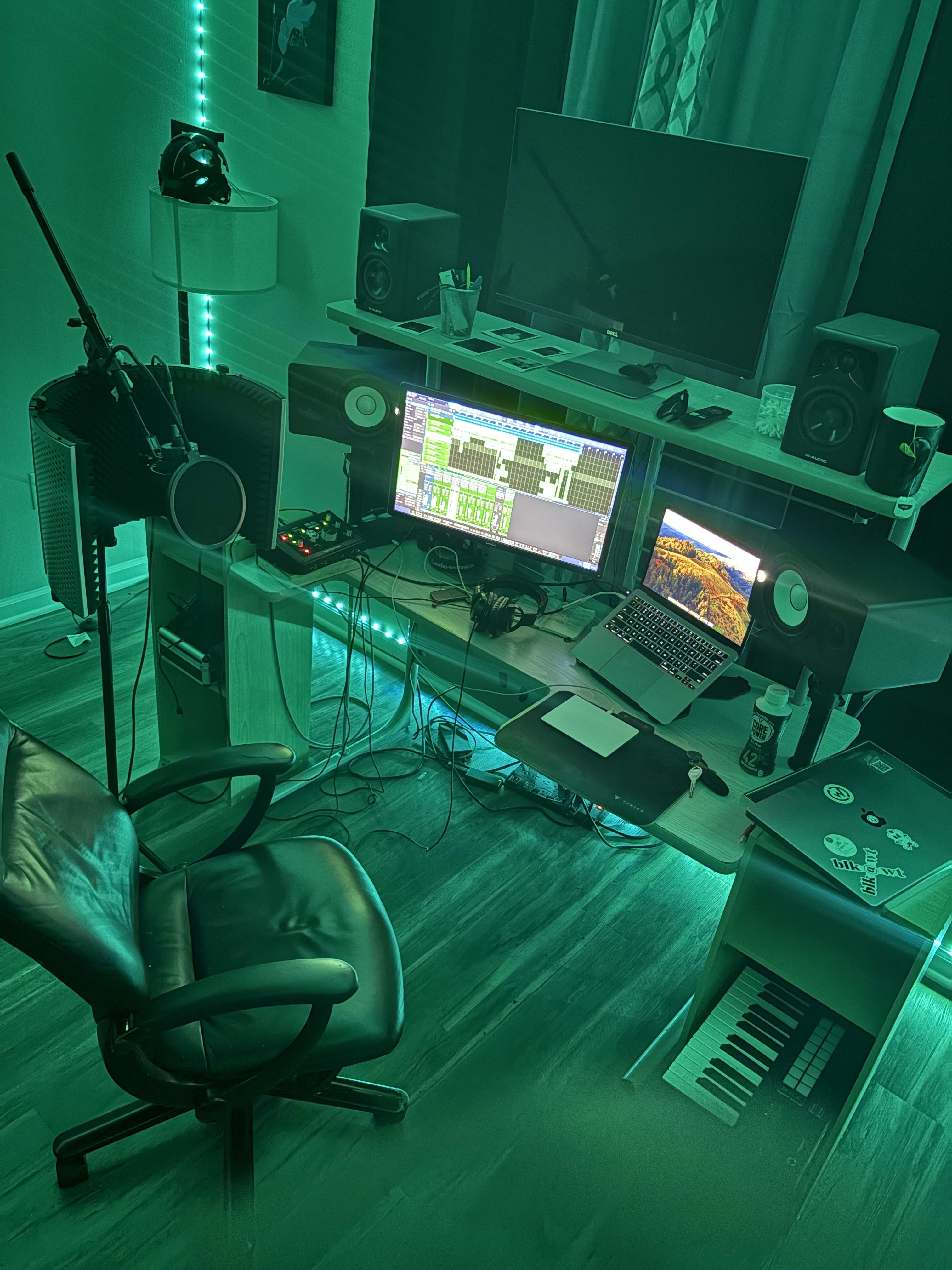Home studio drowned in green LED - dual screens of session tracks, mic shield, keys below the desk