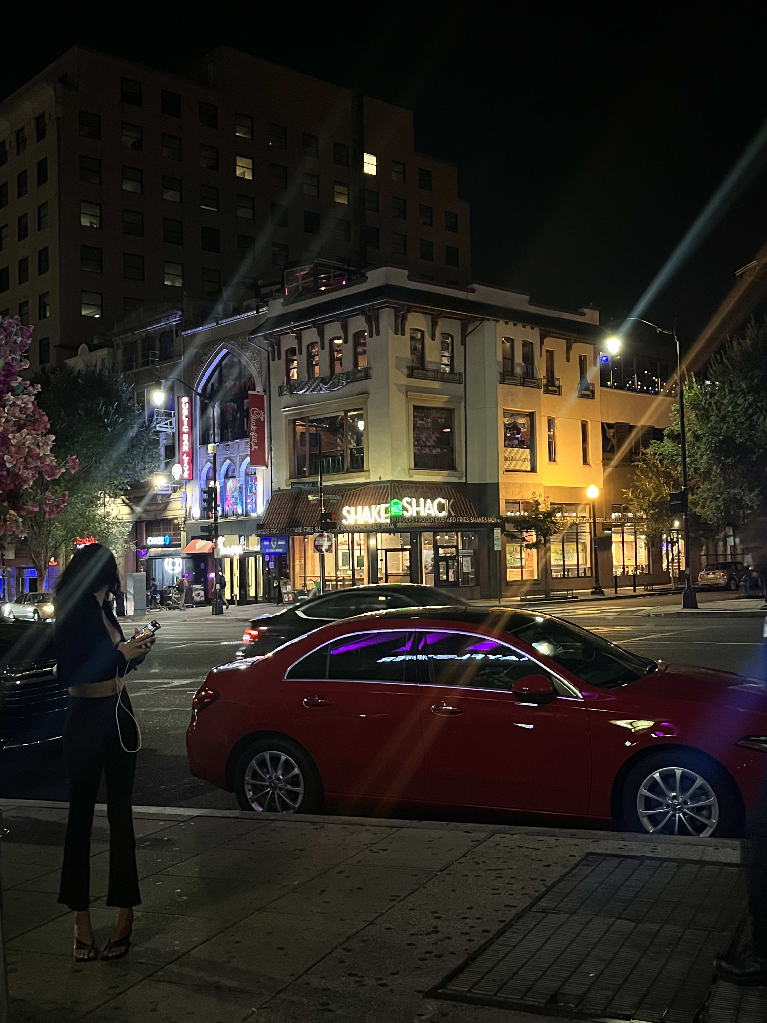 A downtown corner at night - old brick building, neon, a red sedan parked in the glow