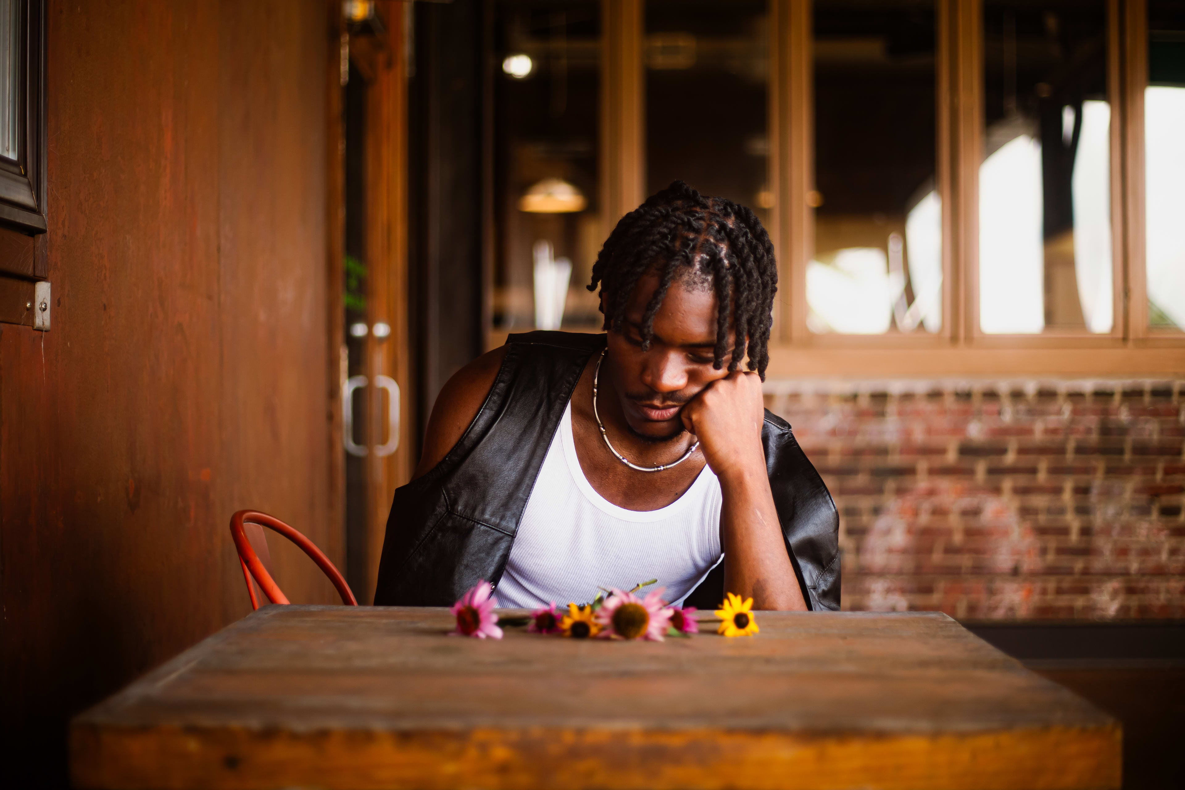 Kama at a wooden table in a leather vest and pearls, chin on hand, wildflowers scattered before him