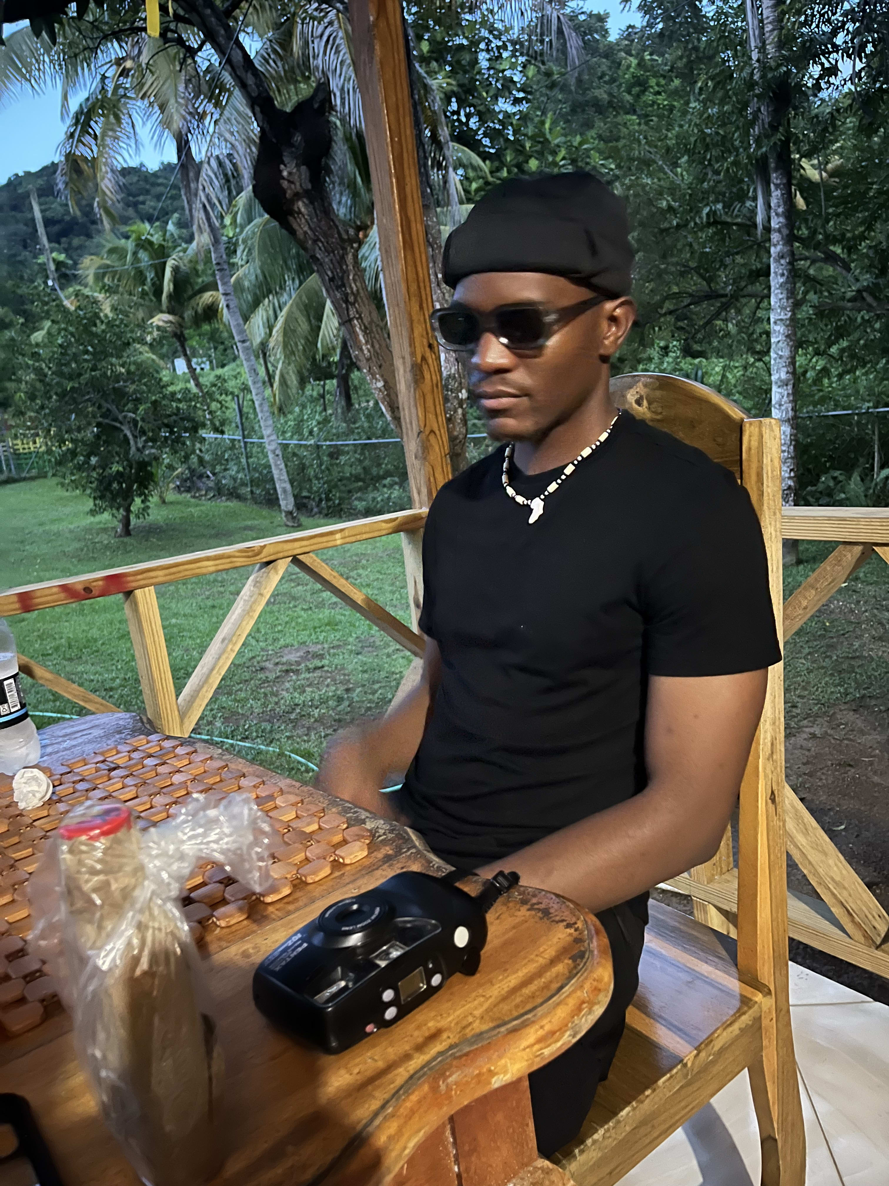 Jamaica: seated in a gazebo in all black, beaded necklace and shades, film camera on the table