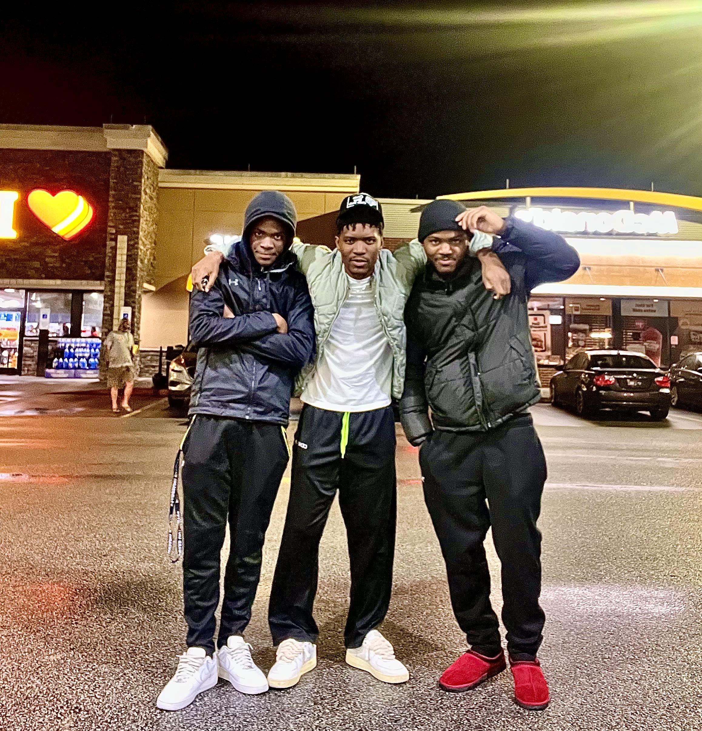 Three friends arm-in-arm at night in a gas-station lot, golden arches glowing behind
