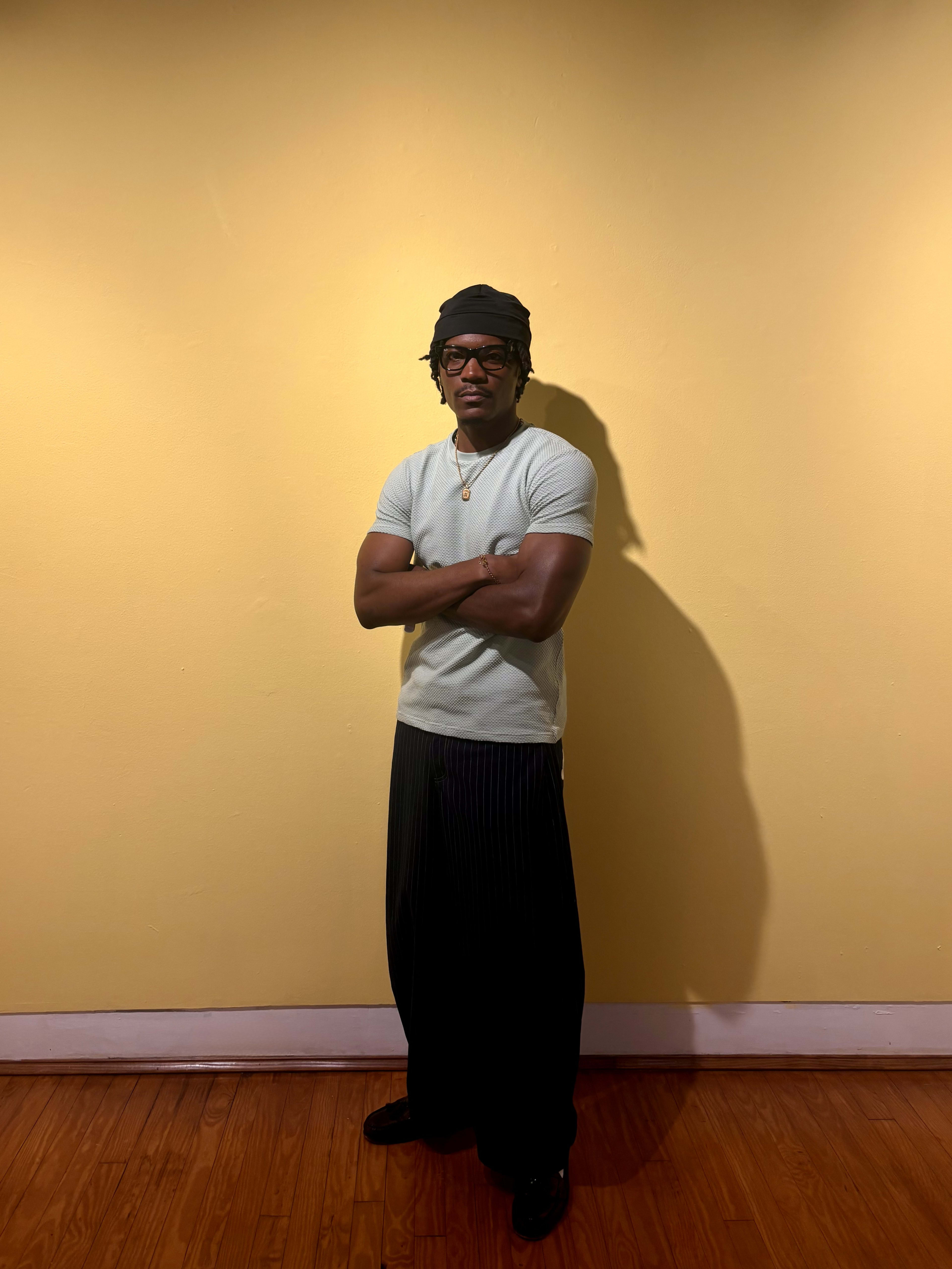 Kama full-length against a yellow wall - beret, glasses, mint tee, wide pinstripes, hard shadow