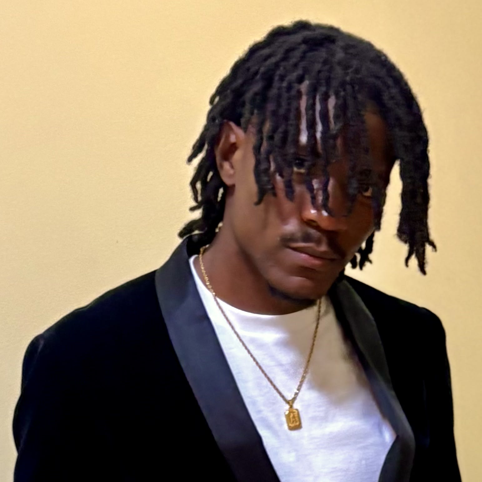 Close crop: locs falling over Kama's face, black velvet tuxedo jacket, gold pendant