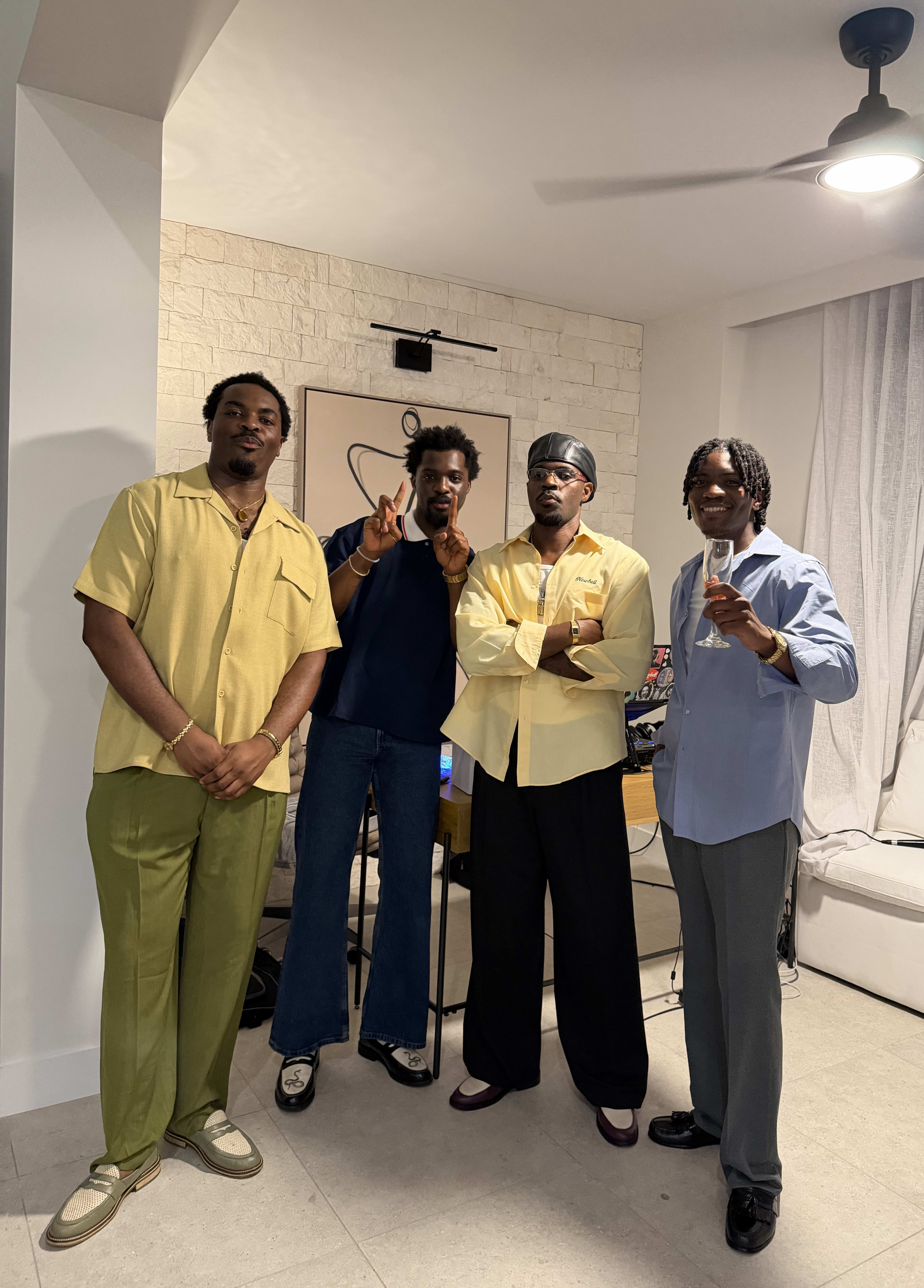 Kama in butter yellow and a durag, arms crossed among three friends dressed for a night in