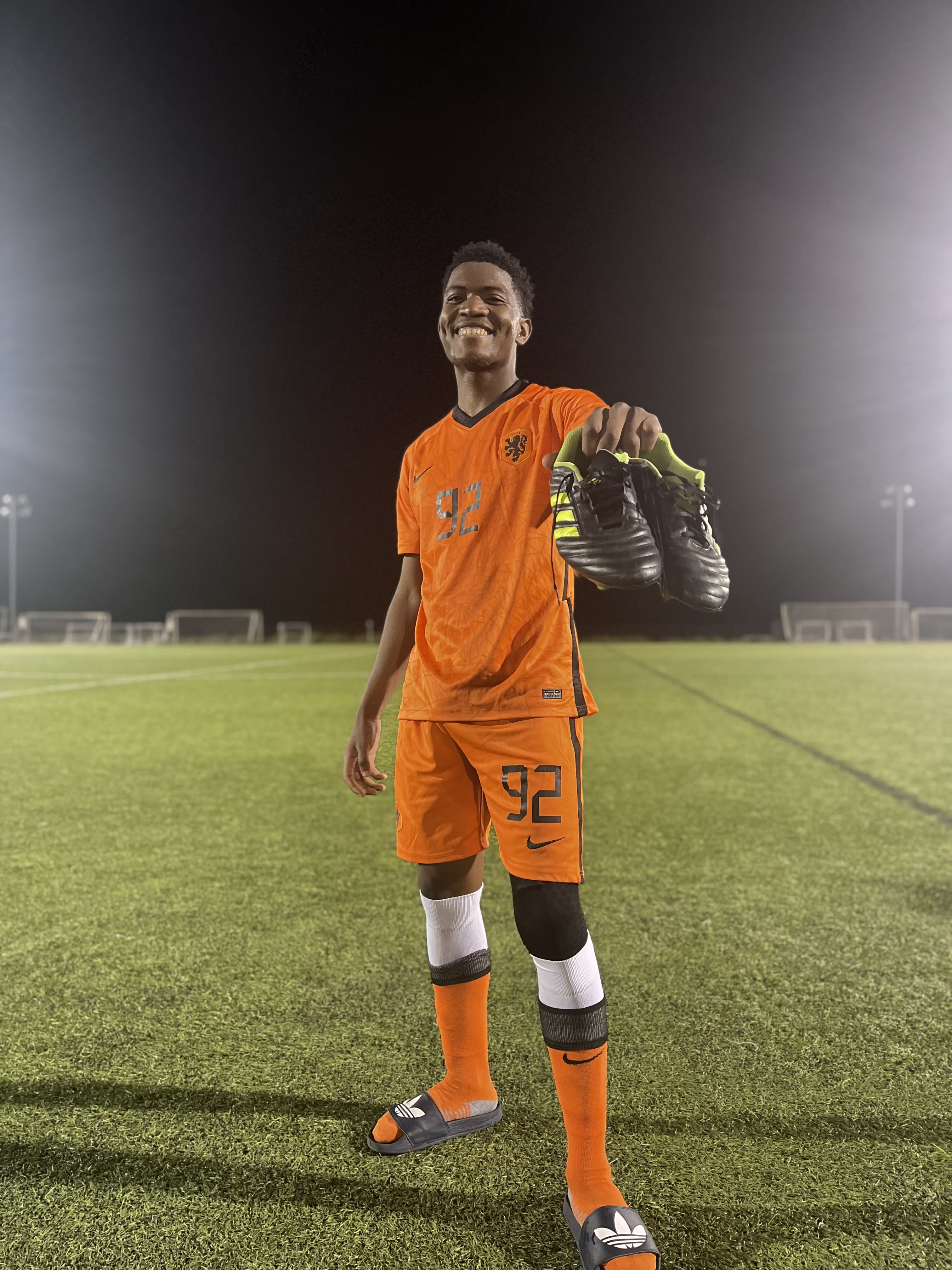 Kama grinning in an orange number 92 kit under the floodlights, cleats held up in one hand