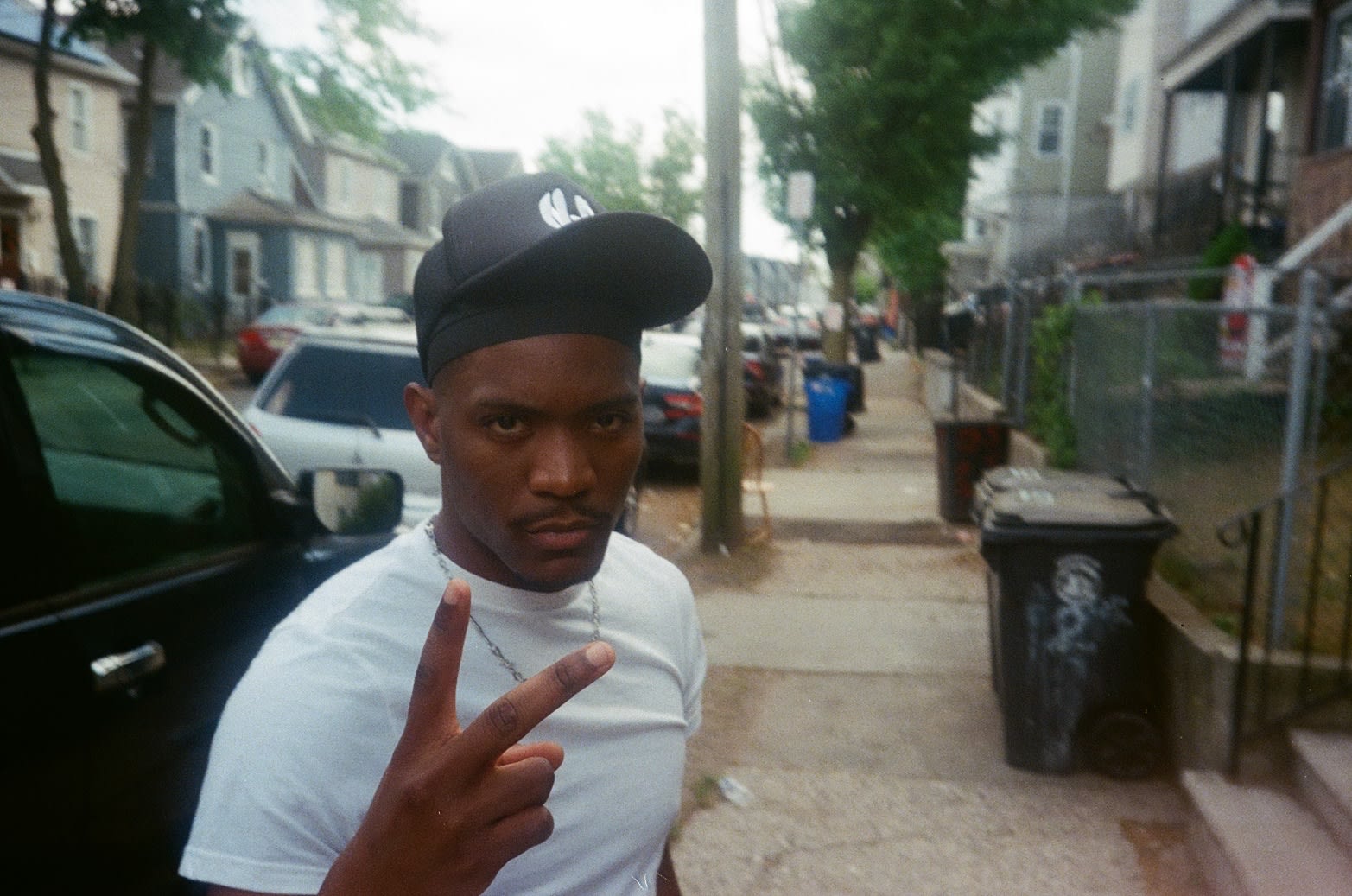 Shot on film: Kama in a white tee and black cap throwing a peace sign on a residential sidewalk