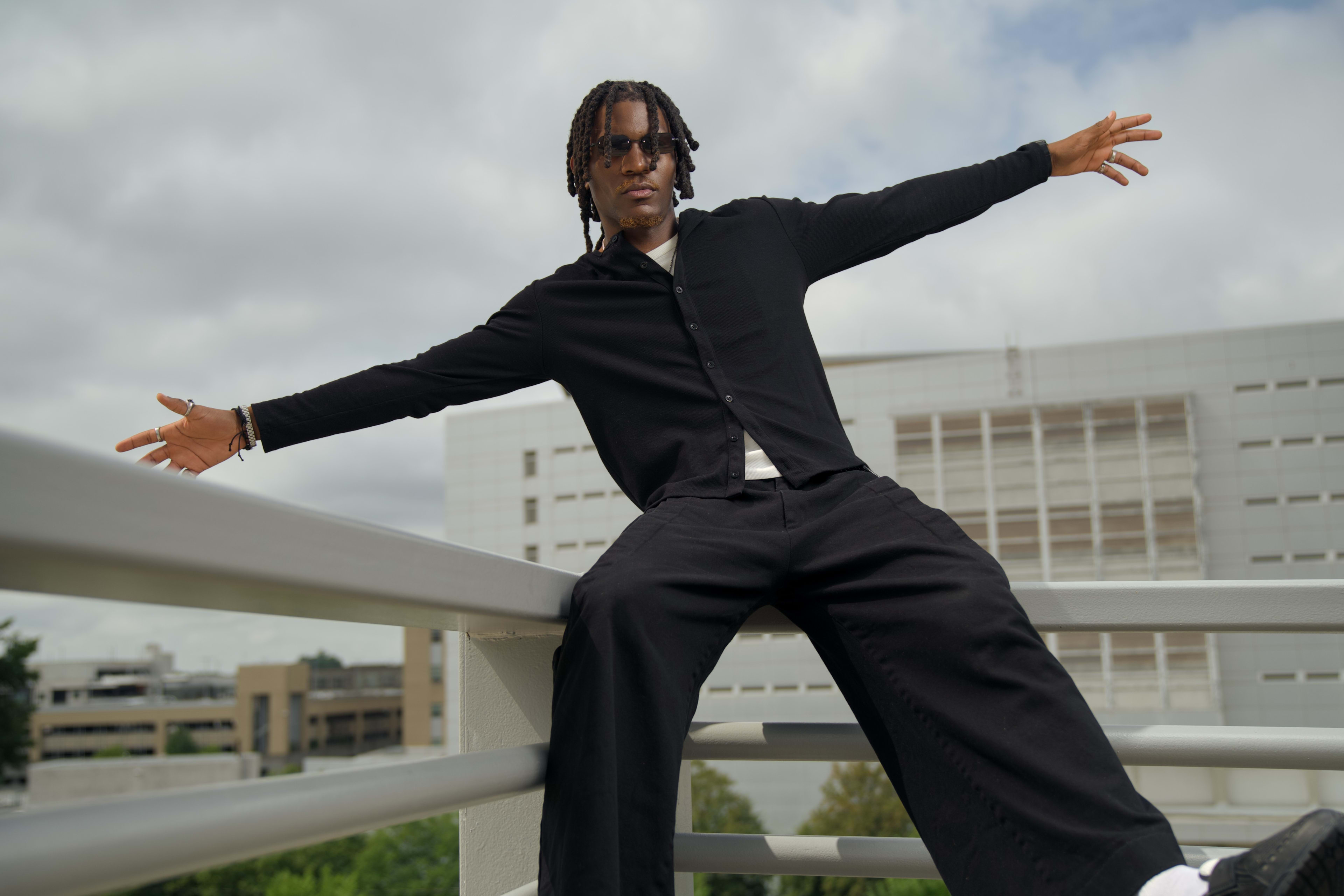 Kama in all black on a white rooftop railing, arms spread wide against an overcast sky
