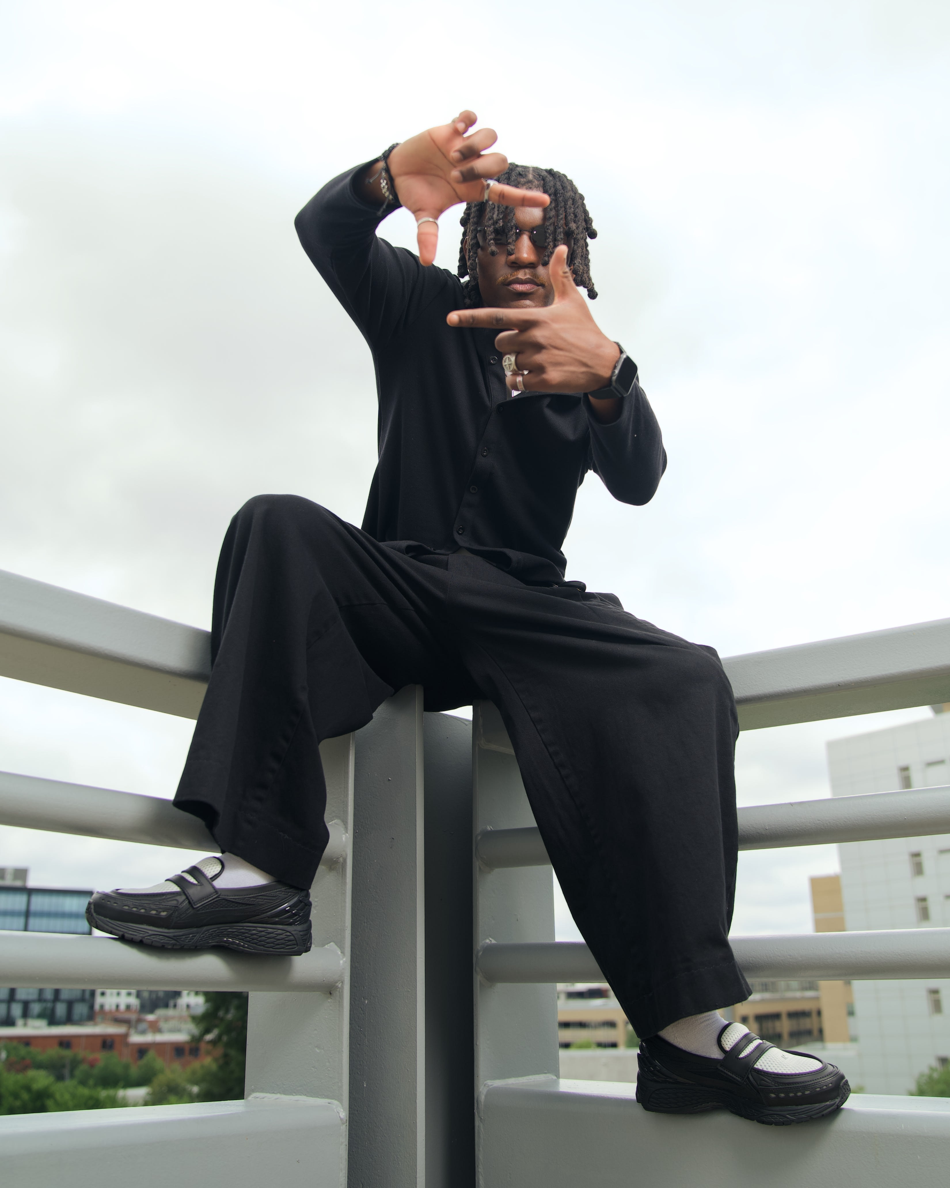 Kama seated on the railing corner, framing the camera between his fingers, all black and loafers