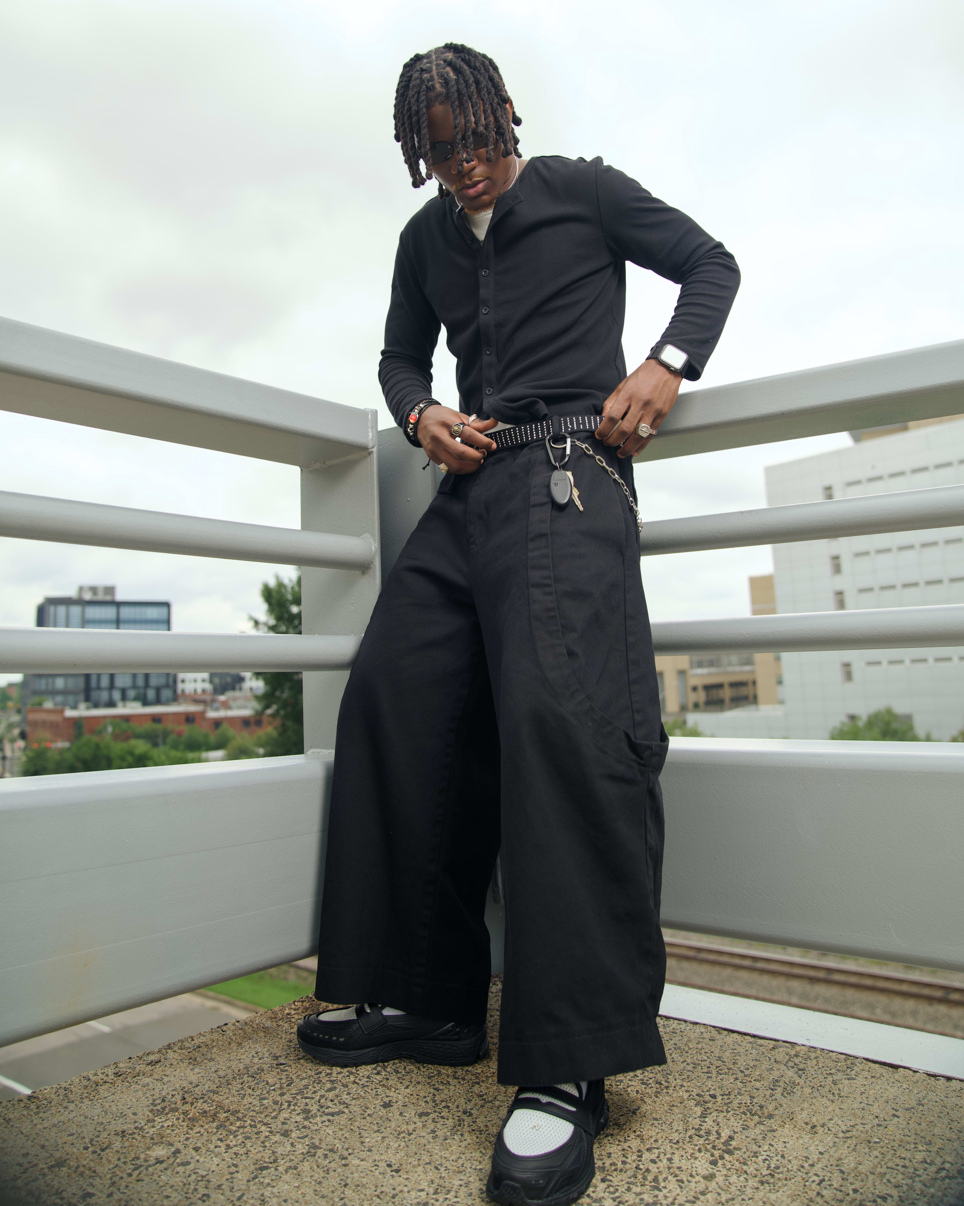 Kama standing at the rooftop rail adjusting a studded belt, wide-leg black trousers draping