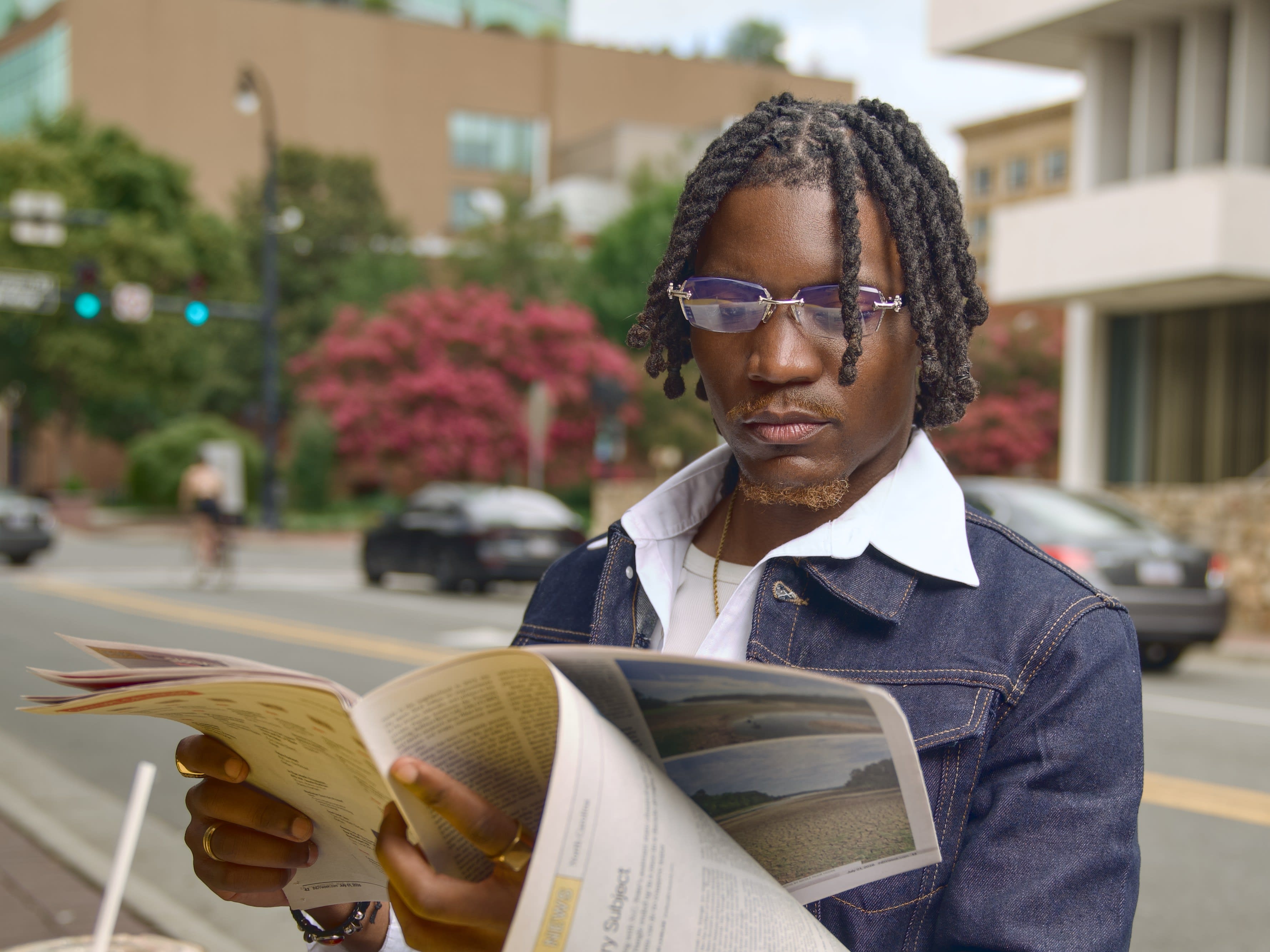 Kama reading a newspaper on the downtown sidewalk in double denim and tinted glasses