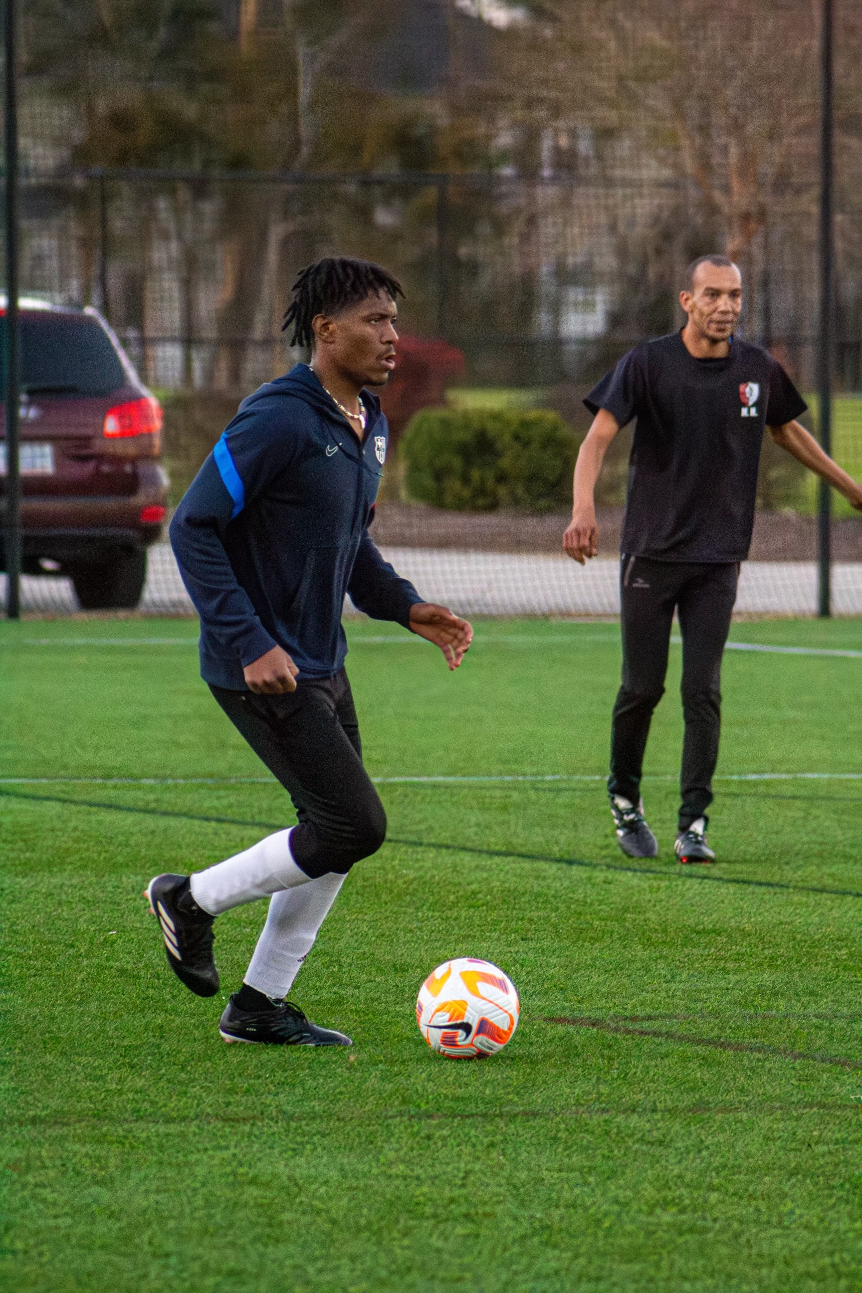 Kama dribbling up the turf in a navy Nike hoodie, eyes up the pitch