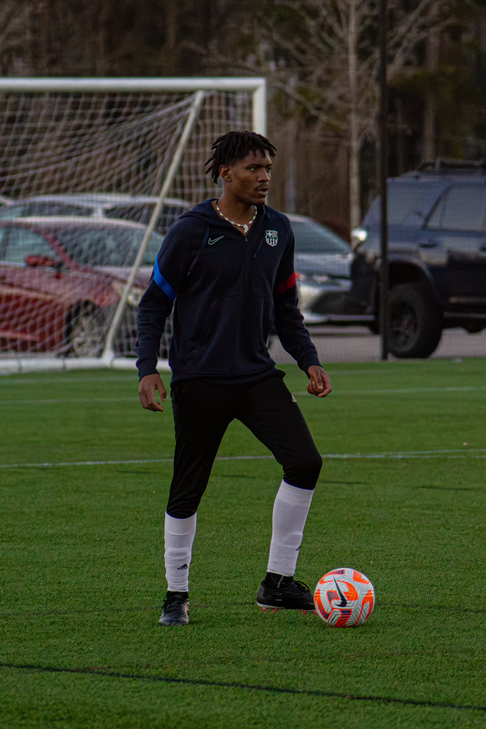 Kama standing over the ball at dusk in a Barcelona zip-up, goal at his back
