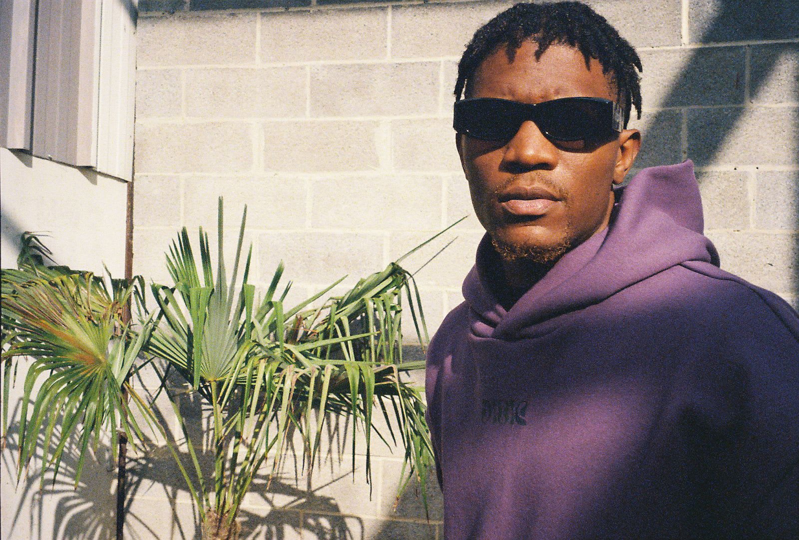 Kama in a purple hoodie and black shades by a cinderblock wall, palms catching the sun