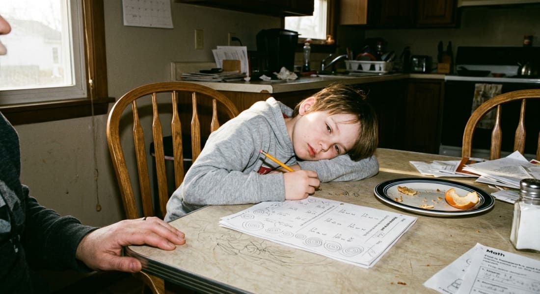 A child at a kitchen table at 4pm, slumped sideways in a chair, pencil in hand, an unfinished worksheet in front of them