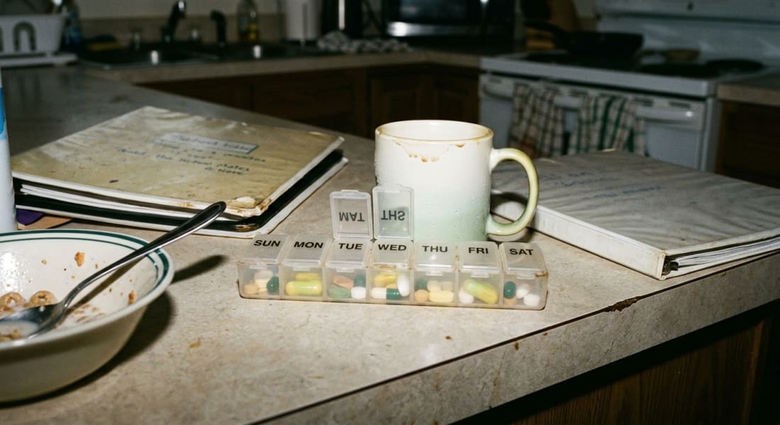 A small plastic pill organizer on a kitchen counter next to a cold coffee mug in morning light