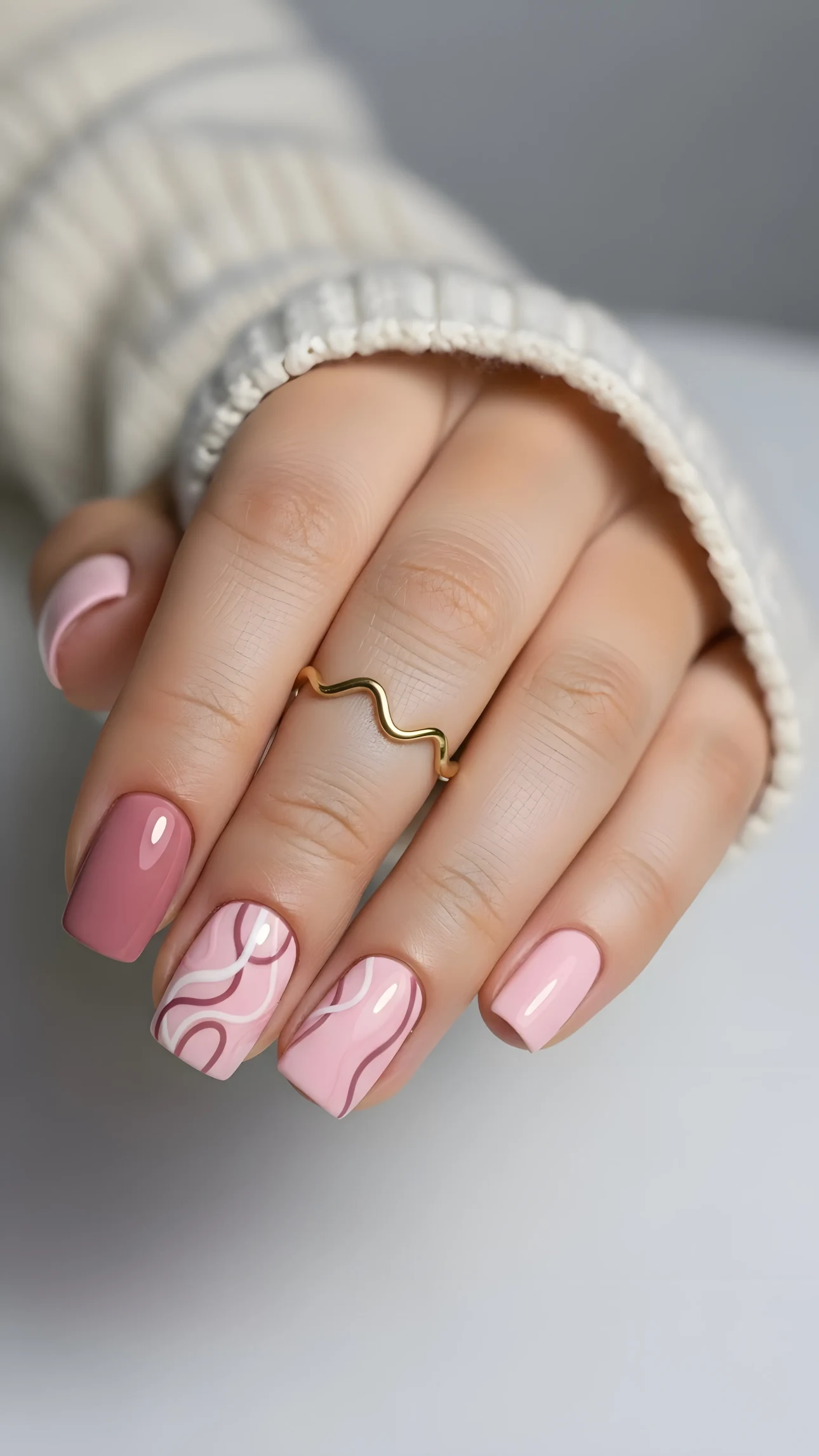 Hand with pink manicure featuring swirl design and gold ring, set against a soft cream sweater background.