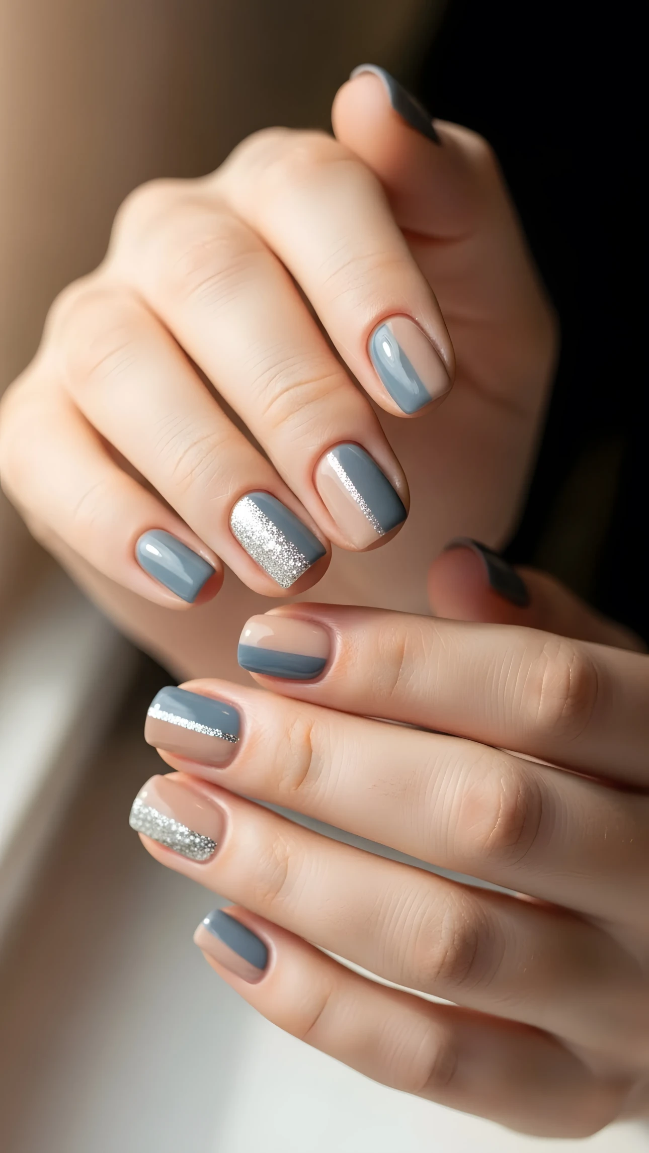 Close-up of manicured hands with gray, nude, and glitter nail art design in soft, diffused lighting.