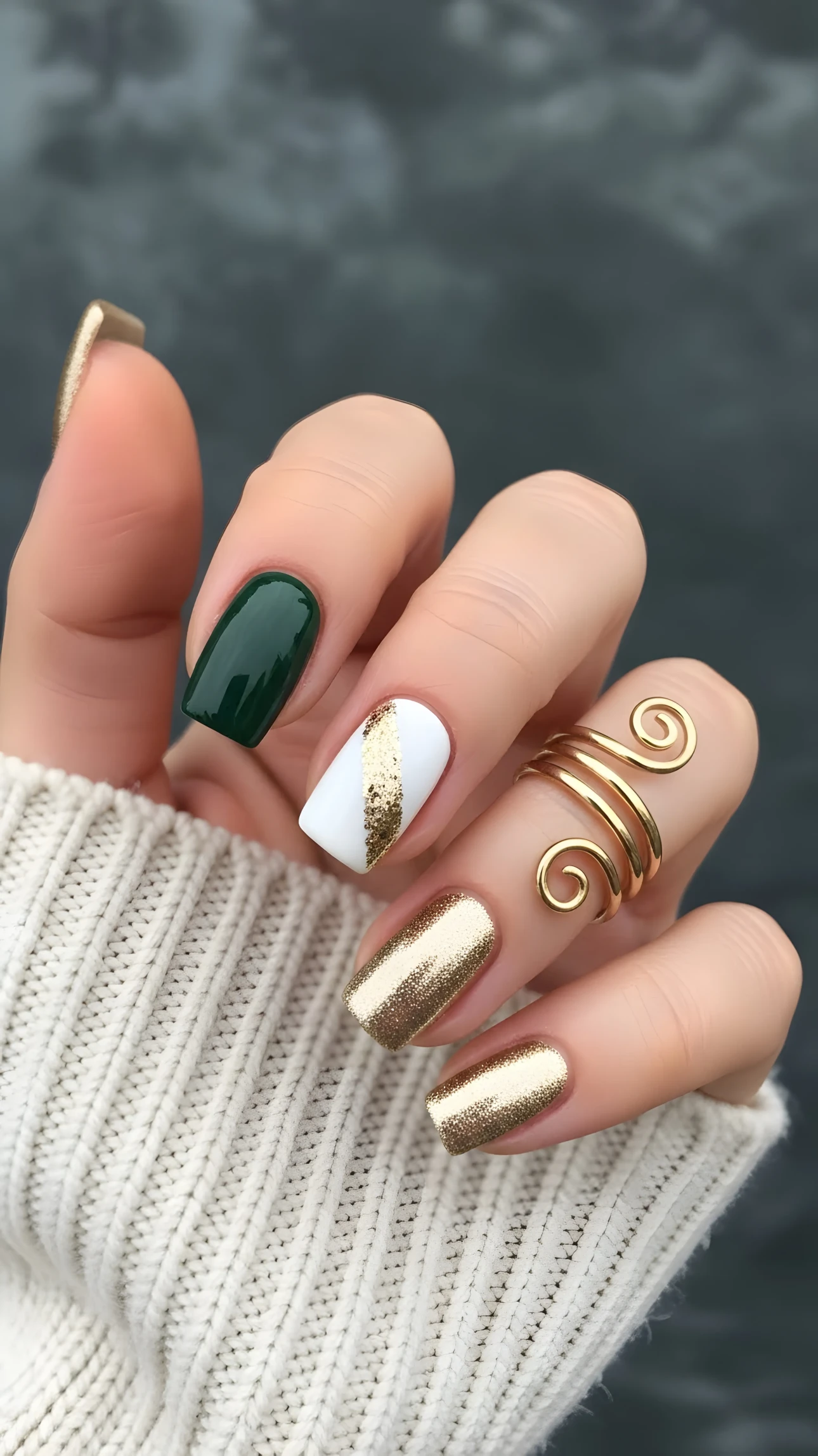 Hand with gold glitter, green, and white nail art and a unique spiral ring, set against a blurred background.