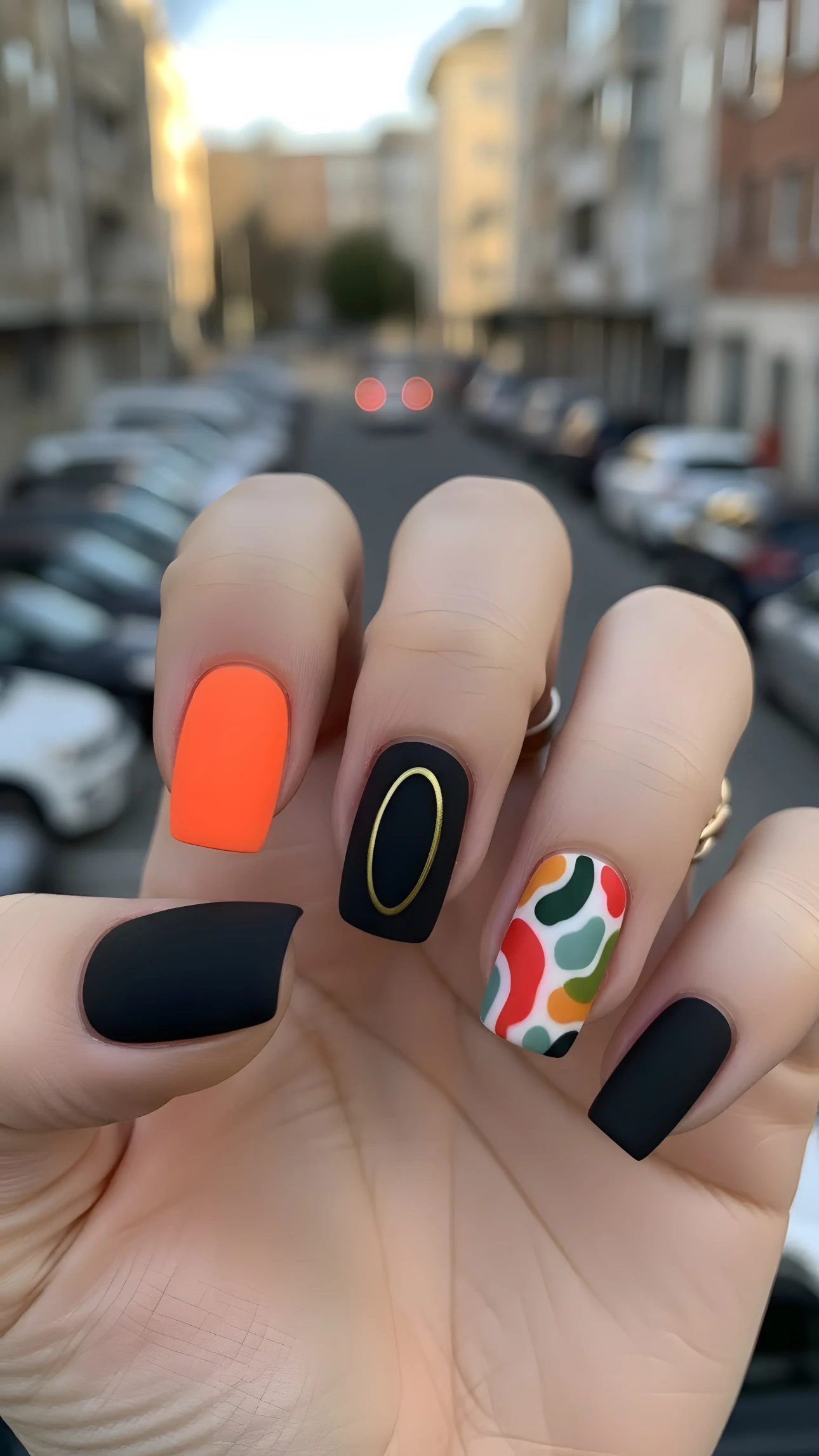 Hand displaying matte nail art with geometric designs, featuring orange, black, and patterned nails in soft outdoor light.