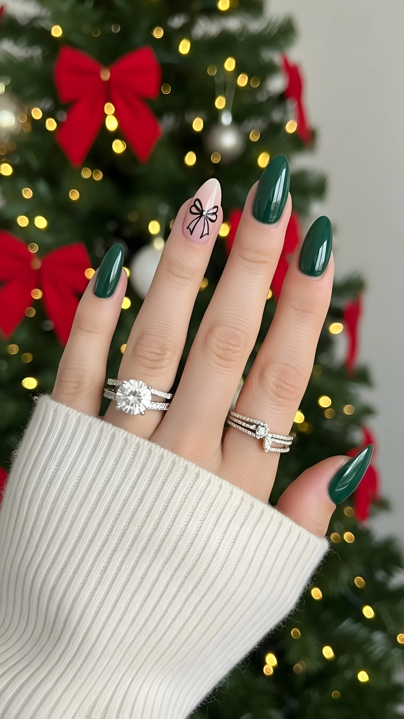 Hand with emerald green nails and diamond rings against a blurred Christmas tree background, showcasing holiday elegance.