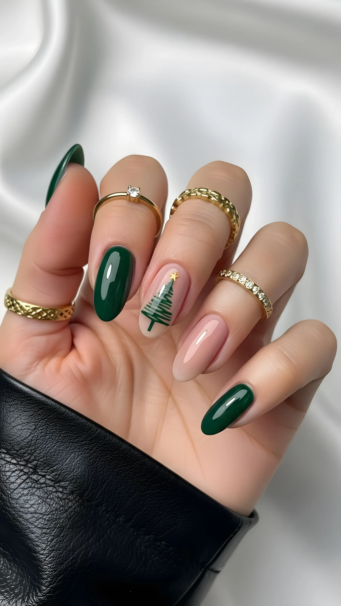 Hand with dark green and Christmas tree nail art, wearing gold rings, against a white silk background.