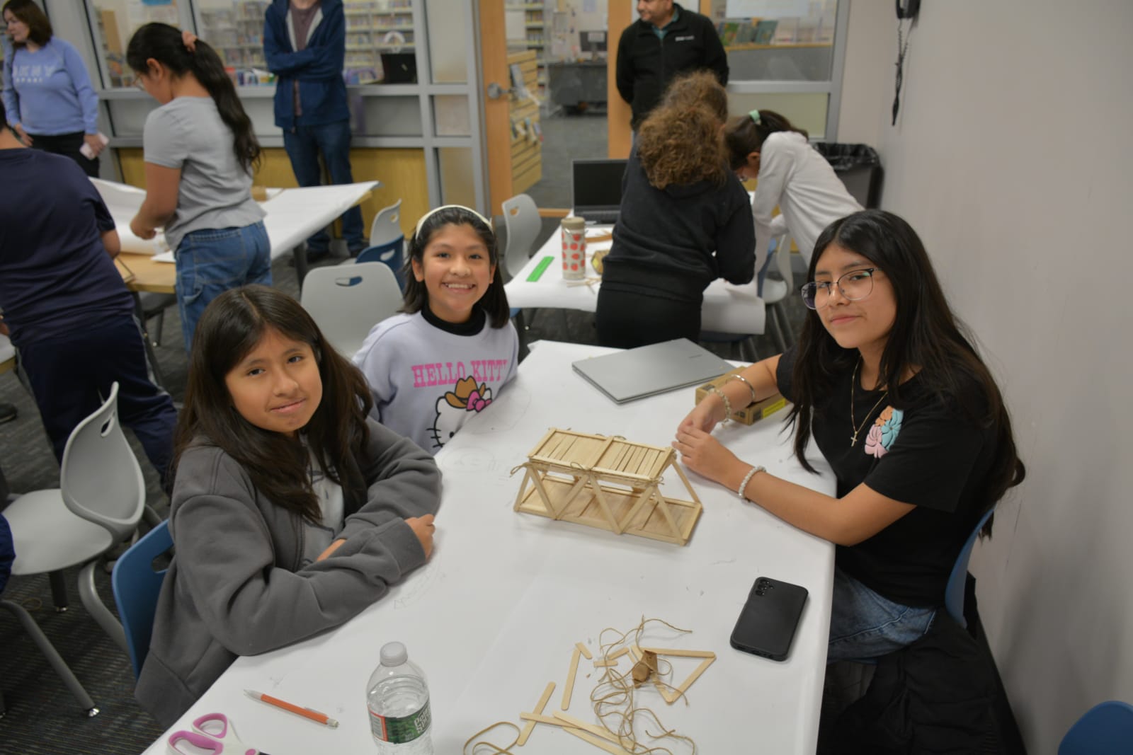 Three students at a library workshop table with the popsicle stick bridge they built