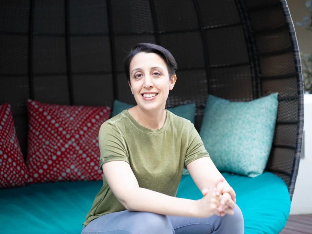 A person with short dark hair, wearing a green shirt, sits on a blue cushioned seat with colorful pillows in the background, smiling at the camera, perhaps after an uplifting session with their therapist in Santa Clarita.