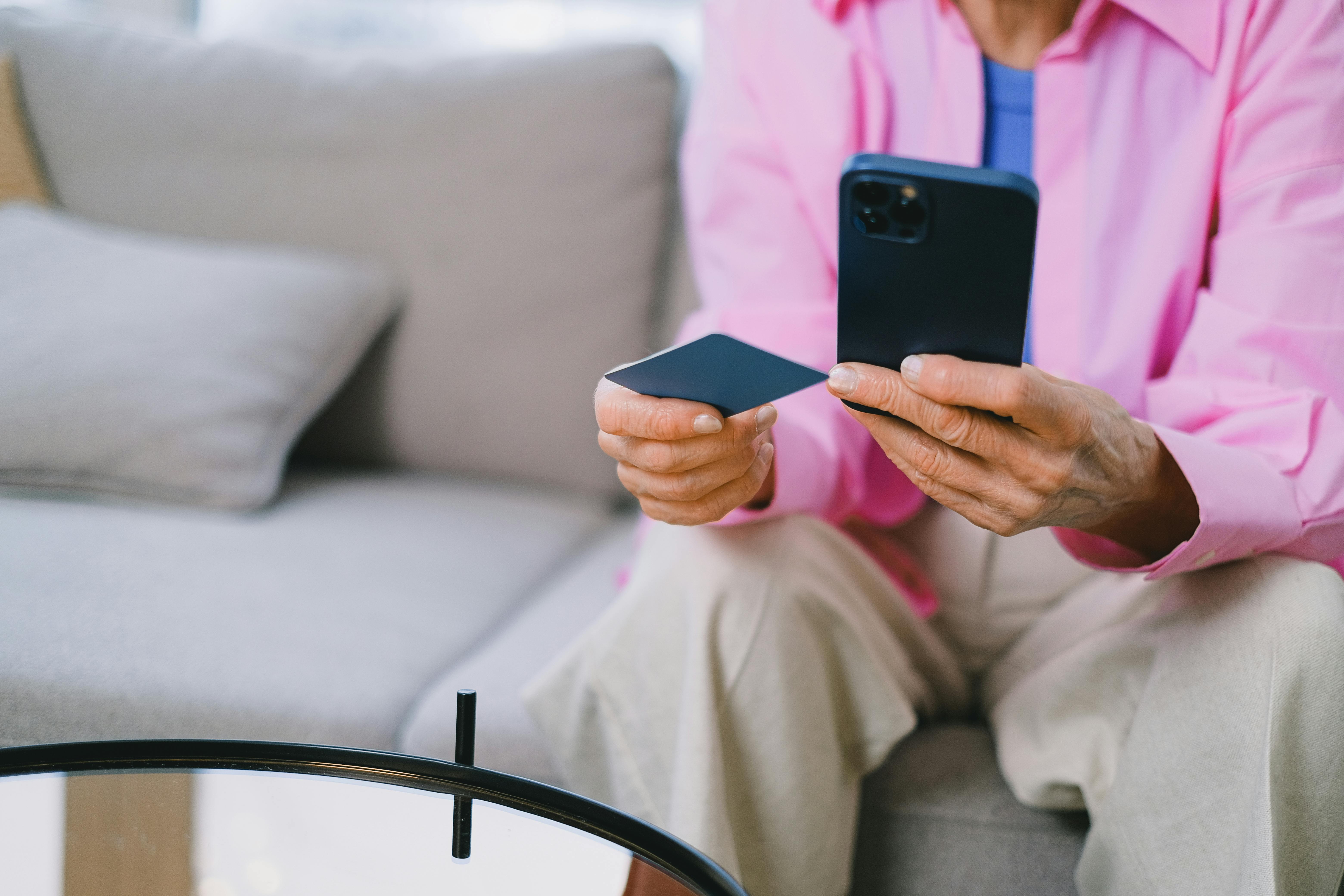 An older person holding a phone in one hand and a card in the other to make an online purchase