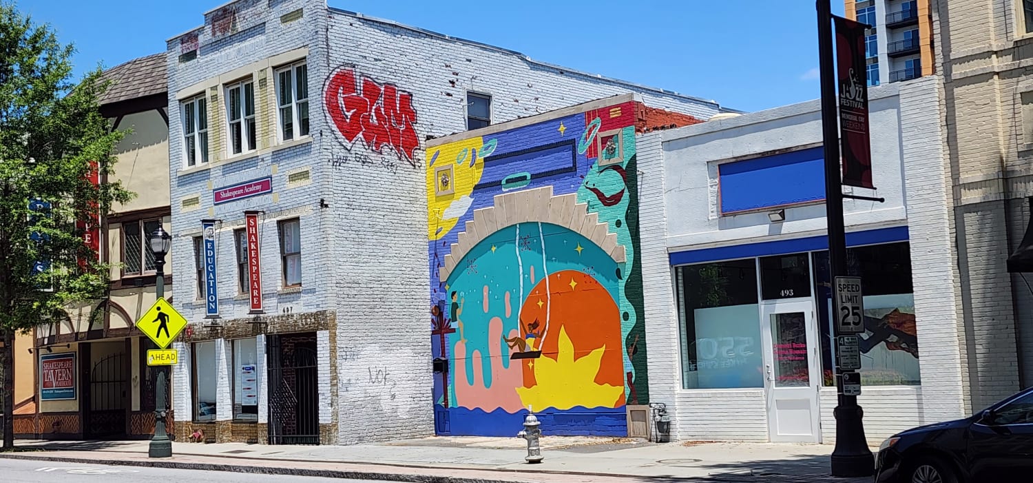 Two buildings. One with a colorful mural of a person on a swing and another bulding with a very large tag with red letters.