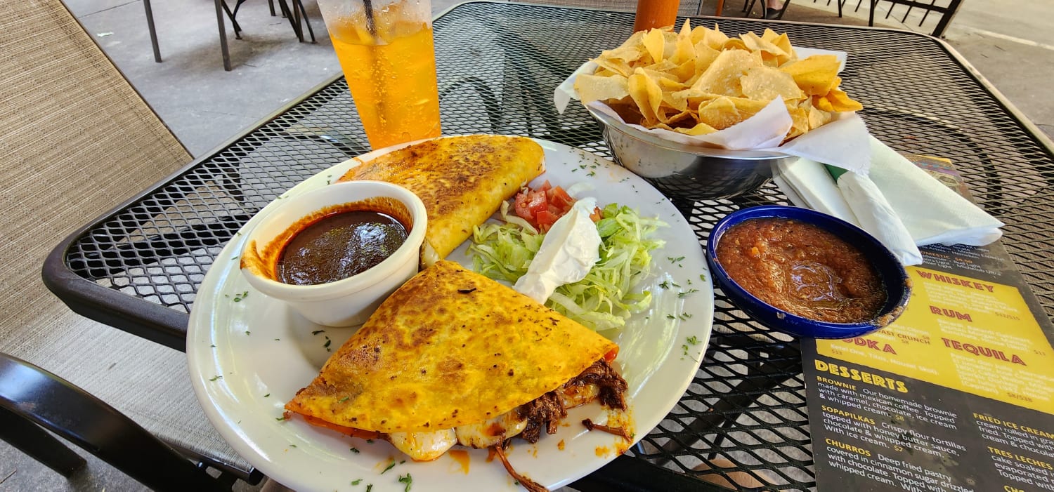 A table with a plate with fajitas tomatoes lettuce sour cream a dipping sauce, salsa, tortilla chips and a glass of orange soda with ice.