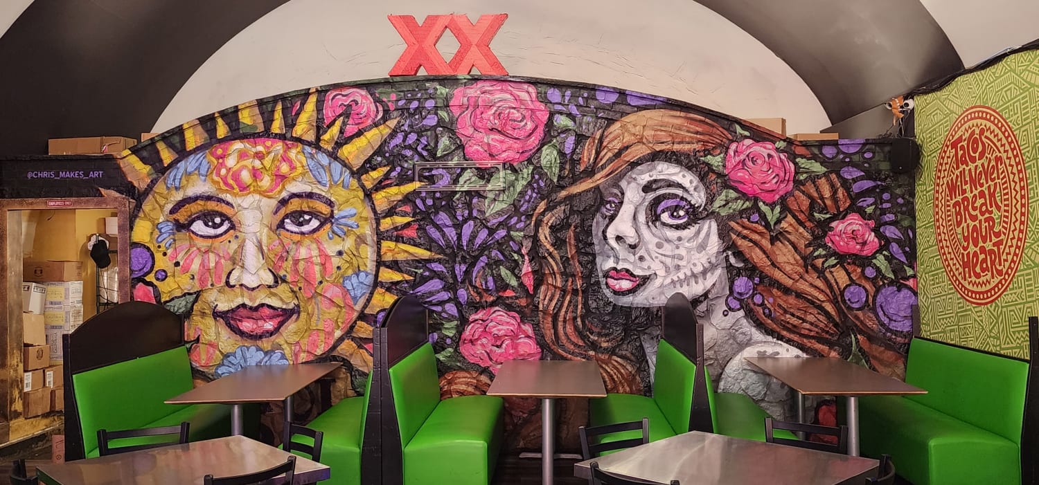 A colorful mural on the back wall of a sun with a colorfully painted face and a woman with their face painted like a skull with roses in their hair and other parts of the mural. There are 2 large red x's on top of the mural. There are some booths with green benches in front of the mural. There is a sign that says tacos will never break your heart to the side.