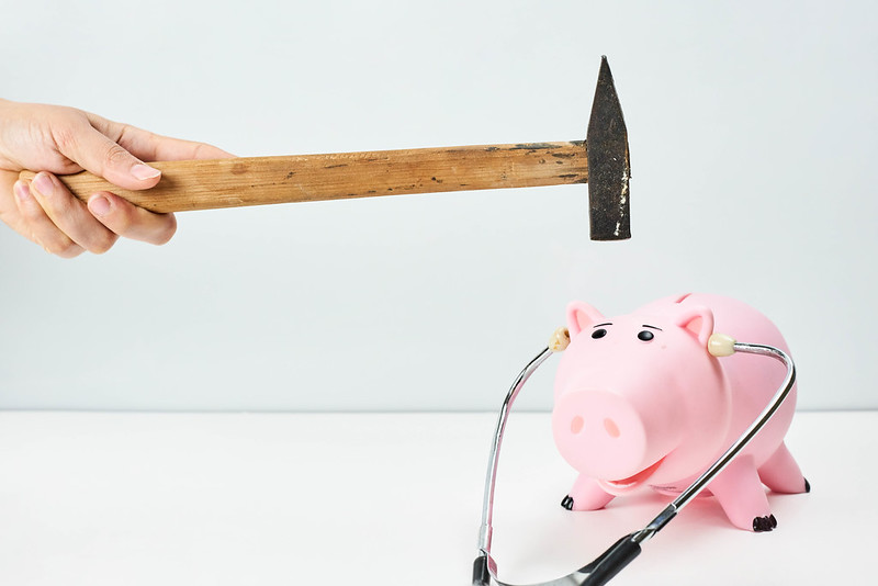 A left hand holds a hammer above a piggy bank with a stethoscope, preparing to smash it to help the hand owner’s small business cash flow