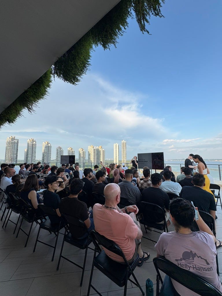 outdoor rooftop event with rows of seated people facing a screen that reads welcome to network school, high-rise buildings and the sea behind