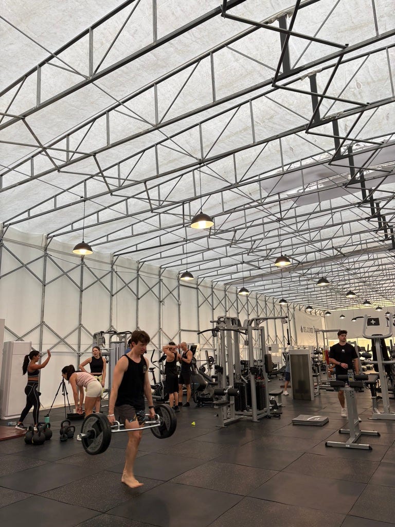 bright indoor gym under a high tent ceiling with metal trusses, people lifting weights and using machines on black rubber flooring