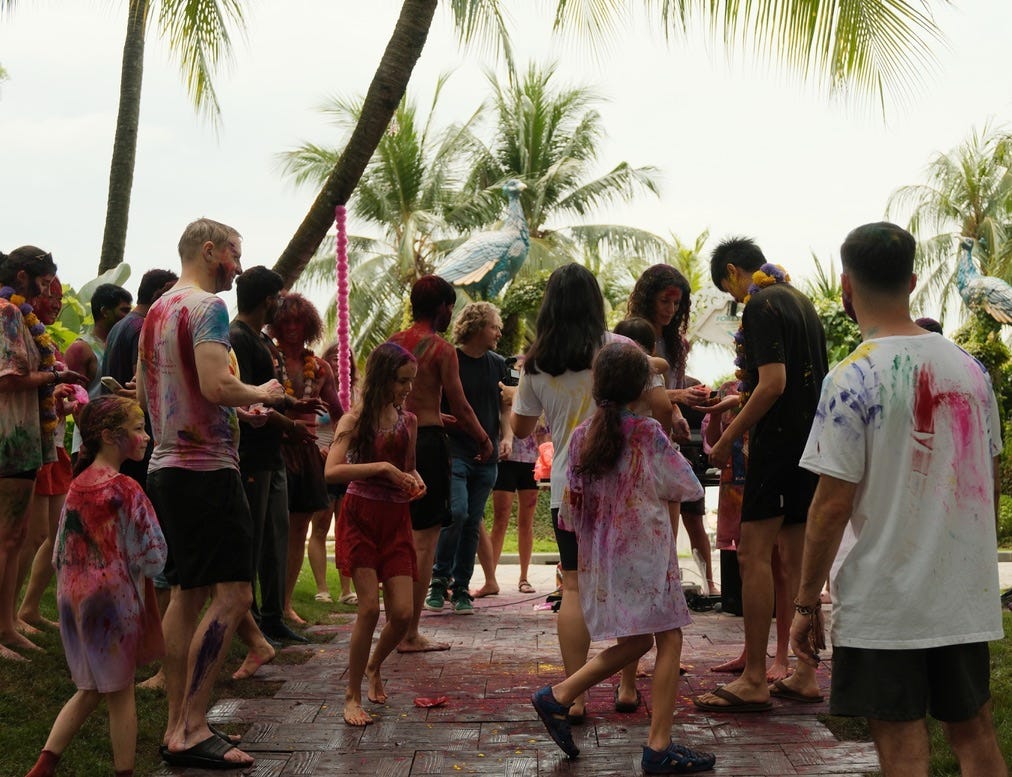 outdoor color powder celebration with adults and children, palm trees, and a large blue peacock statue