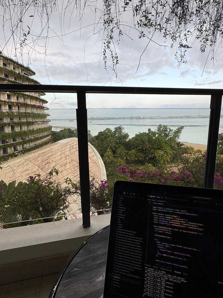 laptop on a balcony table with a code editor open, overlooking tropical buildings, pink flowers, and the water