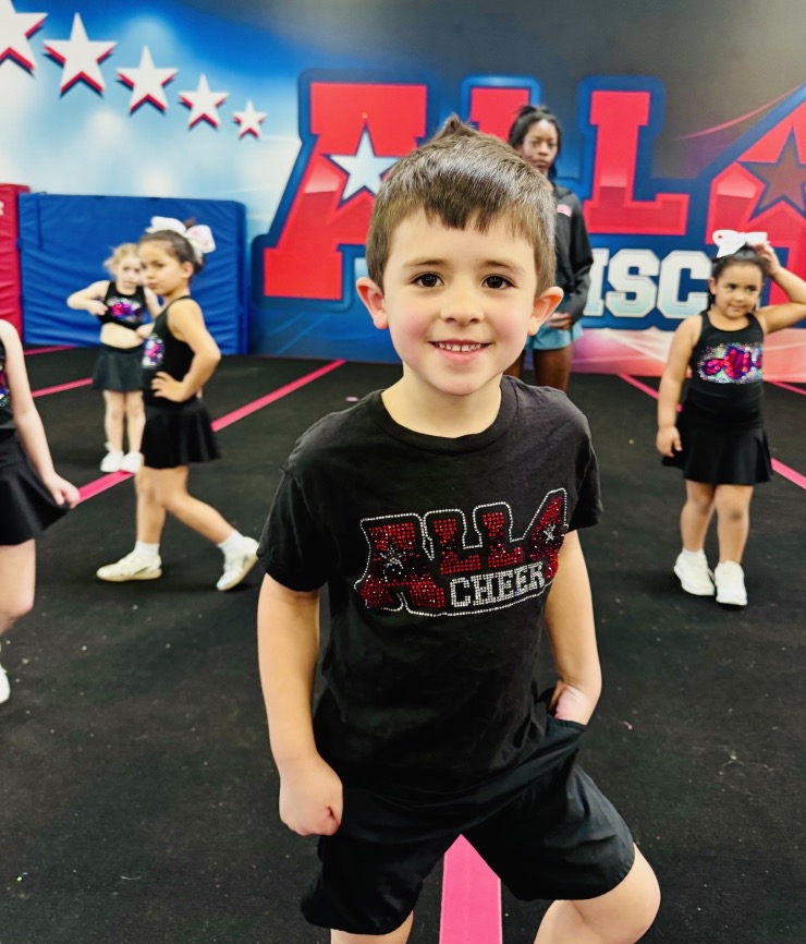 Smiling boy in an All 4 Cheer shirt in front of Tier 1 cheerleaders practicing