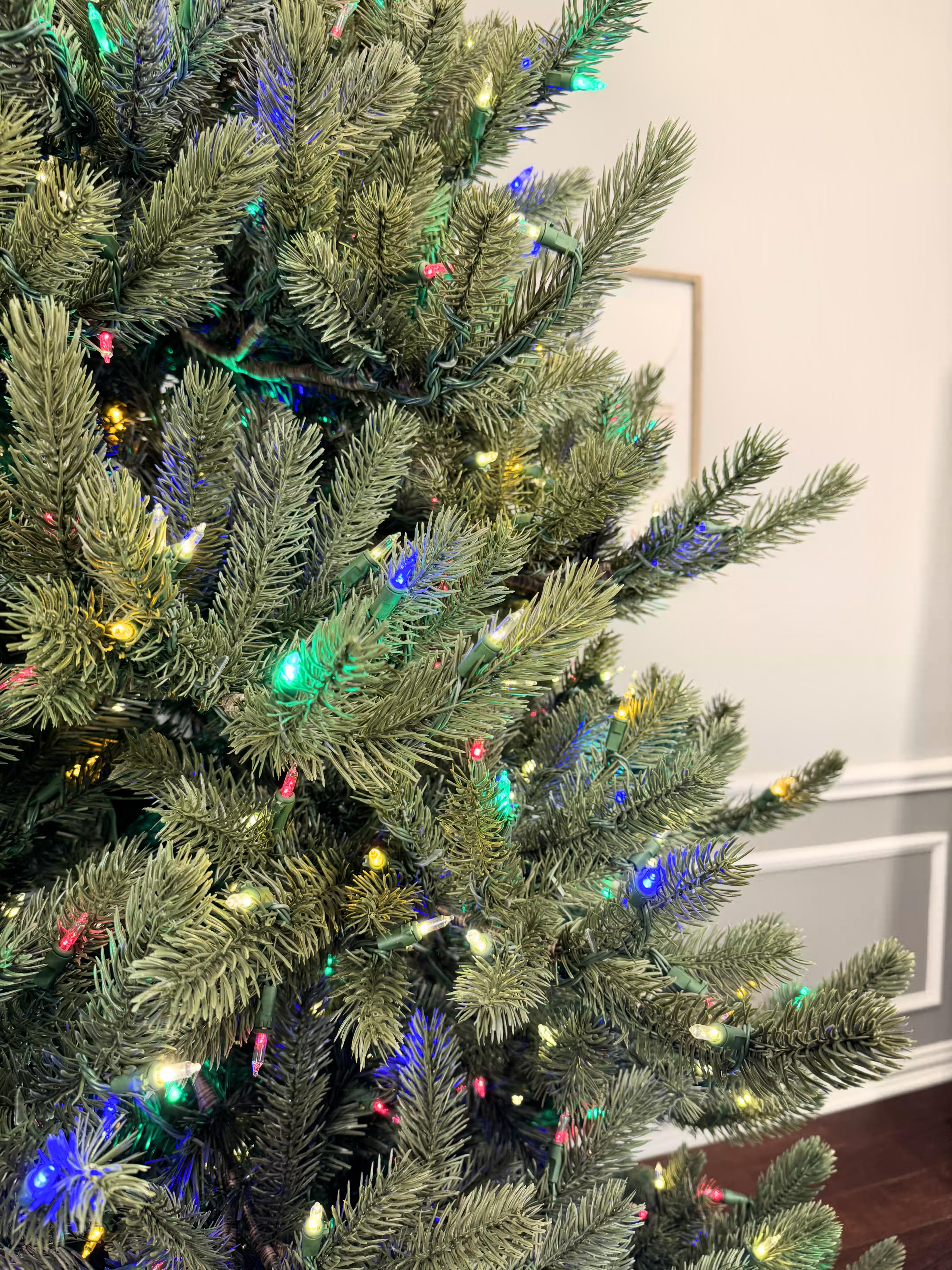 Room lights on with a close up of branch tips with clear+color lights and the wall in the background.