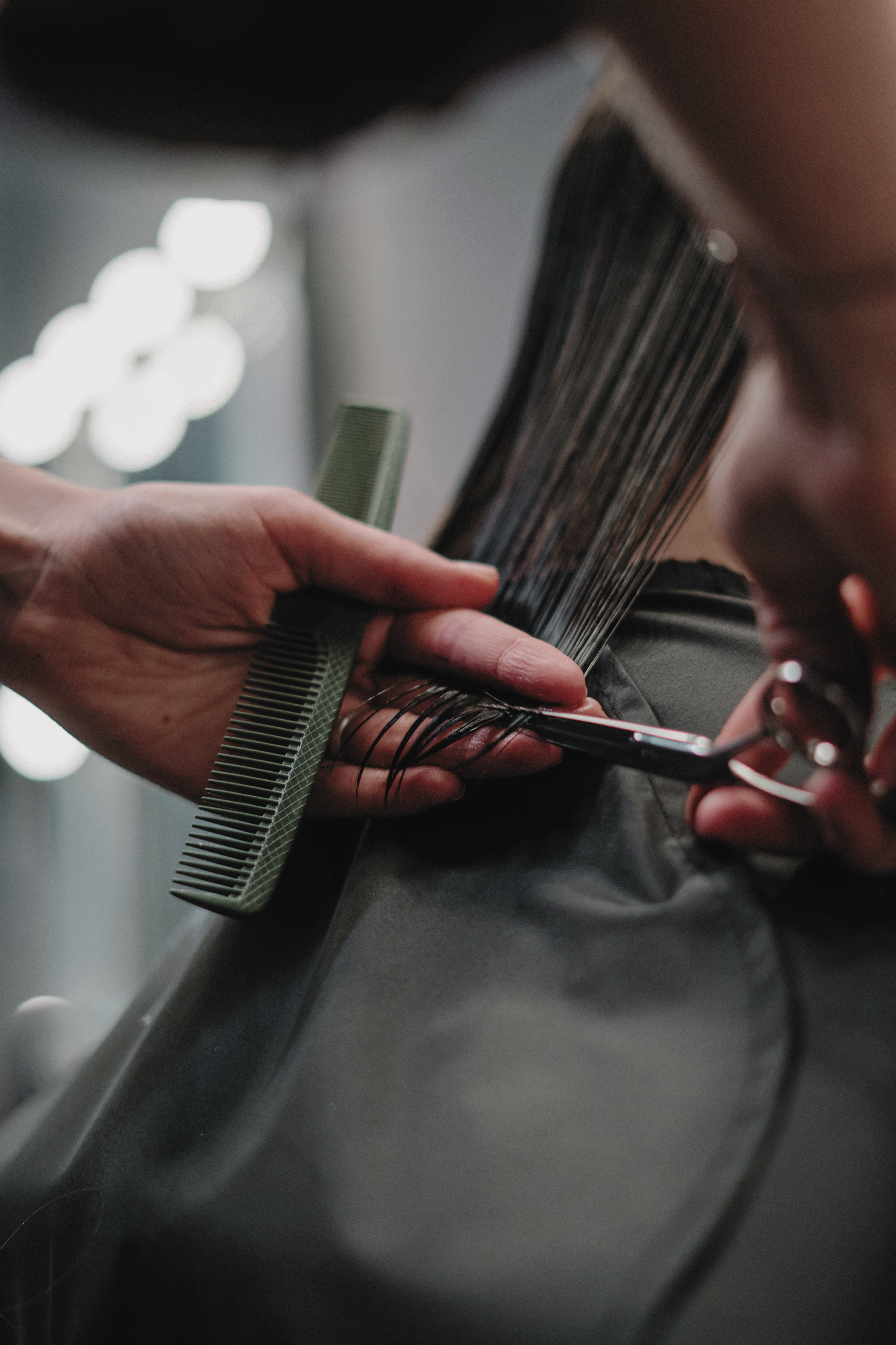 A hairdresser holds a comb in their left hand while their right hand trims someone's medium length dark hair with scissors.