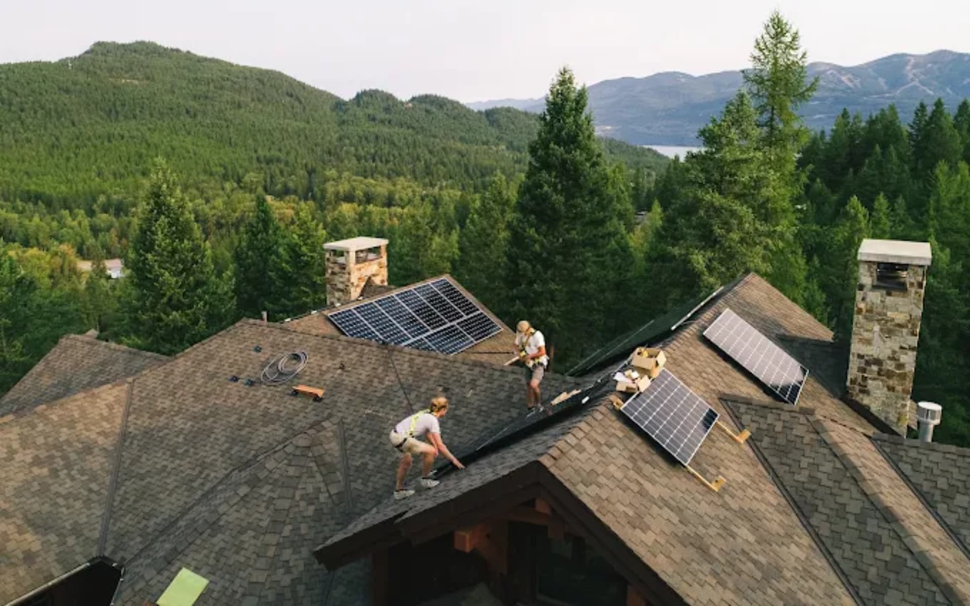 Two installers in harnesses setting solar panels on a shingle roof with stone chimneys, forested hills and a lake beyond