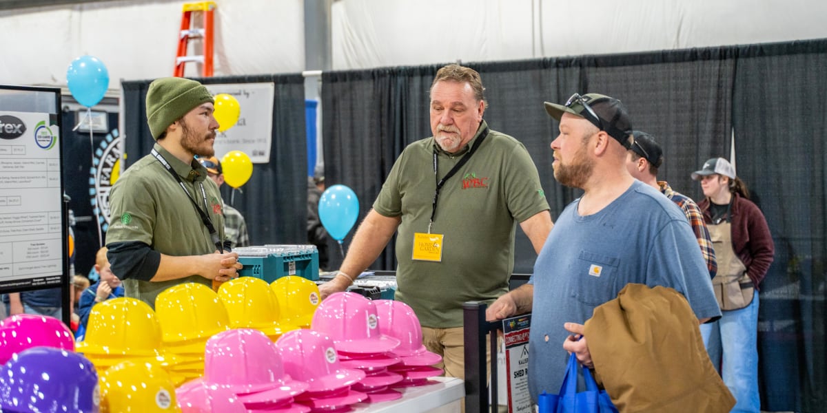 Three men are engaged in conversation at what appears to be a home and garden trade show booth