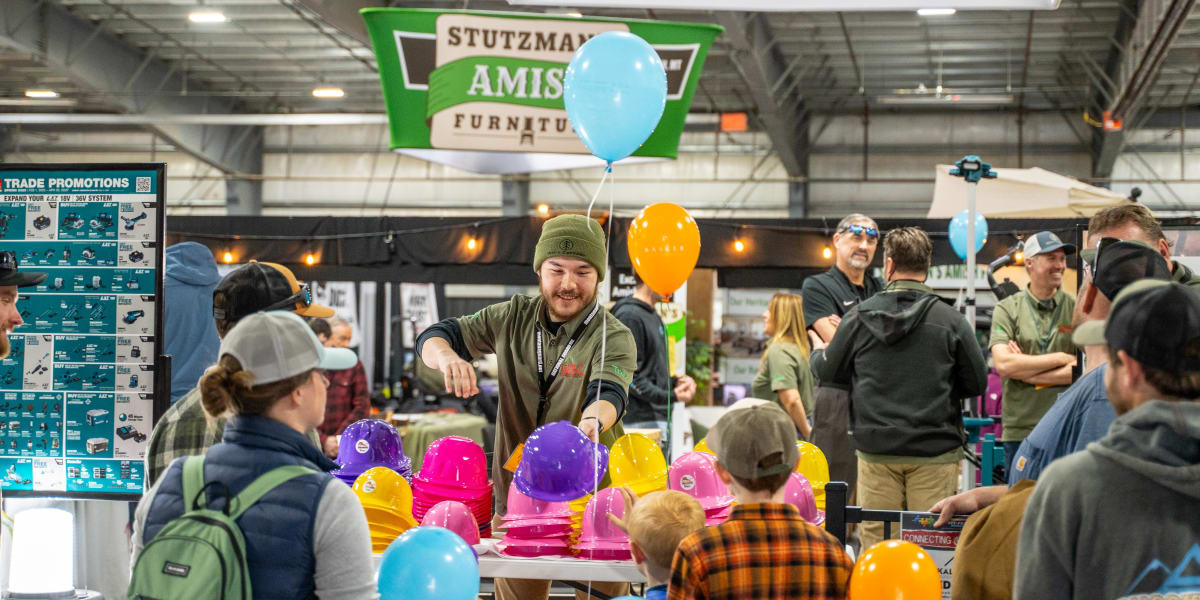 A lively indoor trade show or exhibition scene with a vendor in a green beanie and olive jacket distributing colorful hard ha
