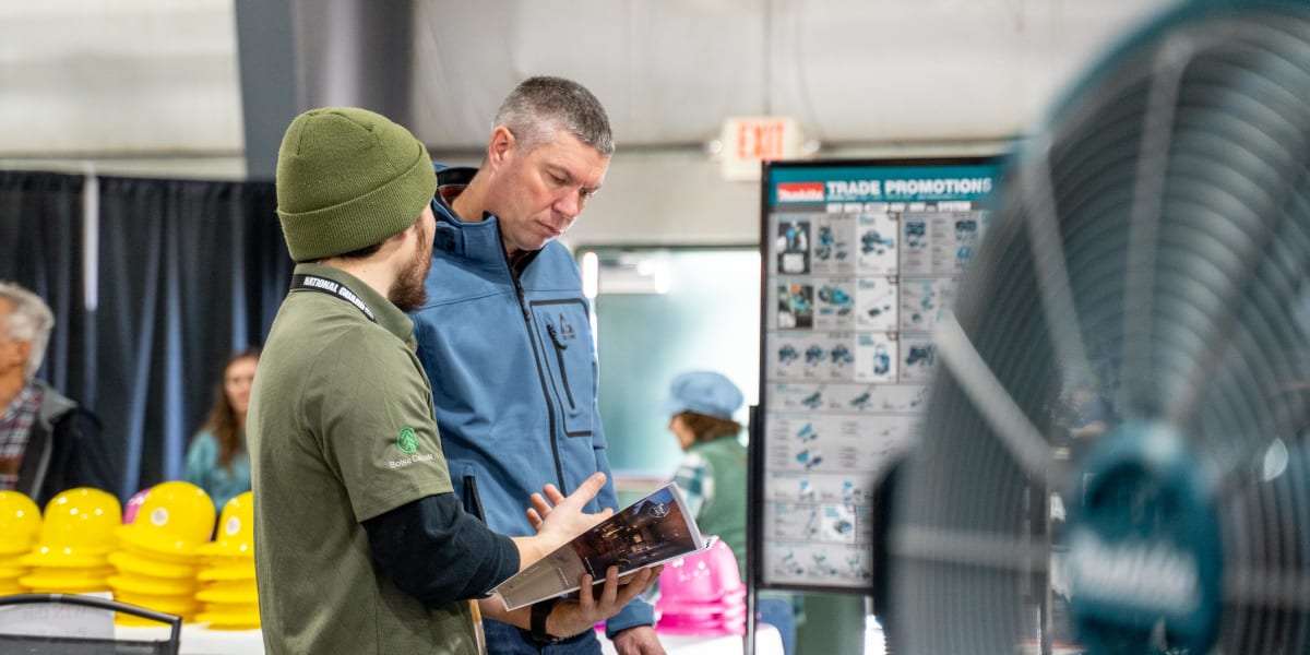 Two men are engaged in conversation at what appears to be a trade show or exhibition, with one wearing a green beanie and mil
