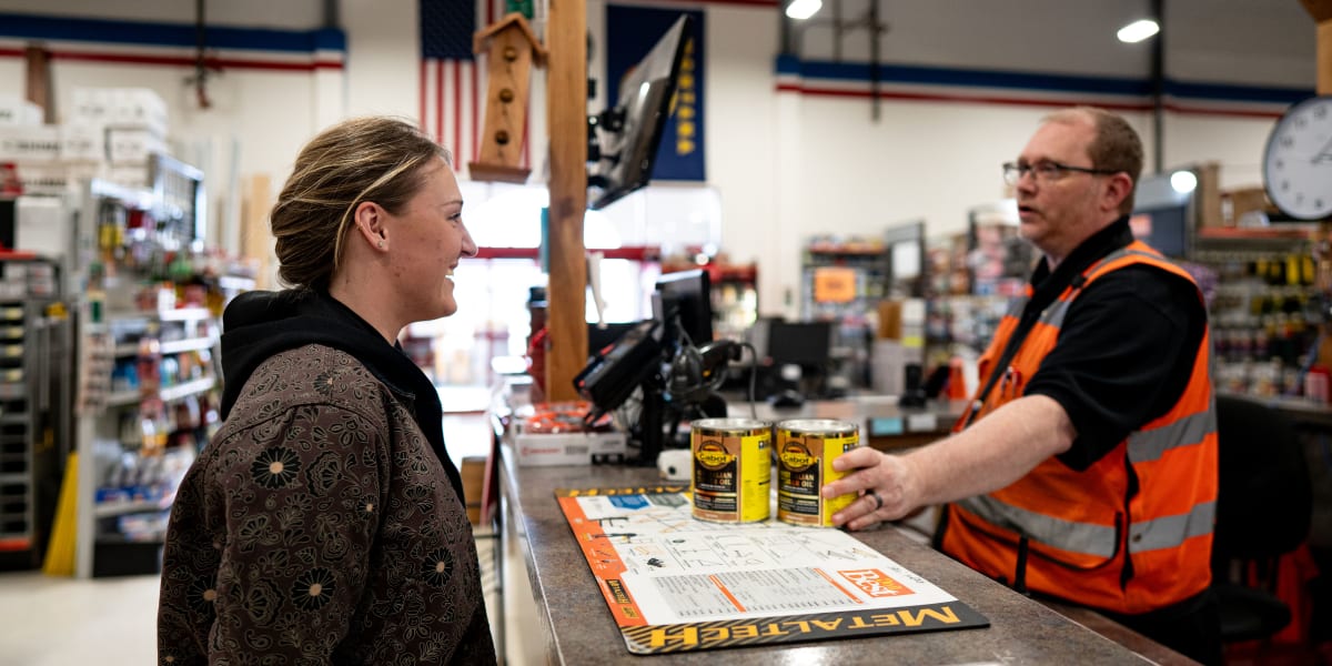 A female customer in a dark patterned jacket is interacting with a male employee wearing an orange safety vest and glasses at