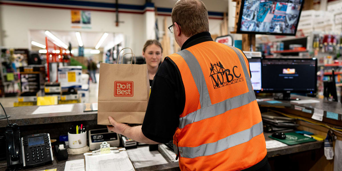A Western Building Center employee wearing an orange safety vest with WBC logo is standing at the customer service counter, h