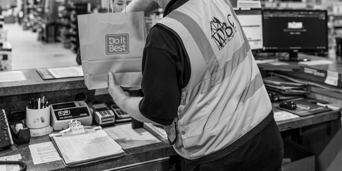 A Western Building Center employee in a company vest is working at a checkout counter, handling a Do it Best branded shopping