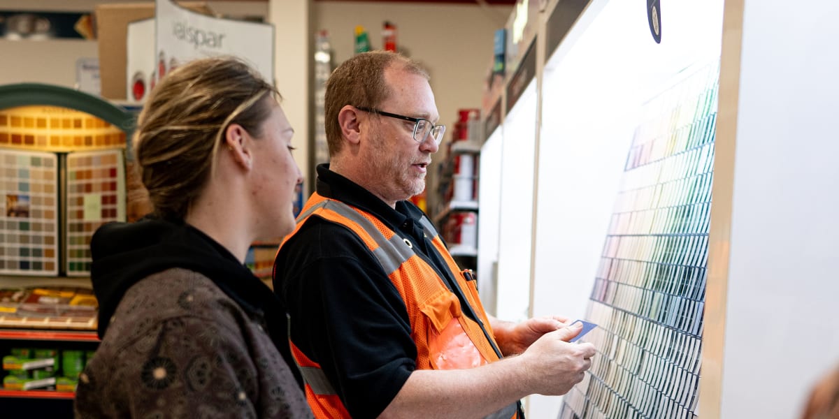 A Western Building Center employee in an orange safety vest and glasses is helping a female customer select paint colors at a