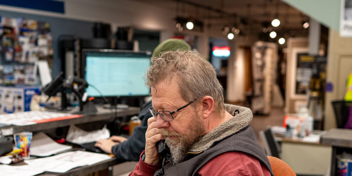 A bearded employee wearing glasses, a red shirt, and high-visibility safety vest sits at a desk talking on a corded telephone