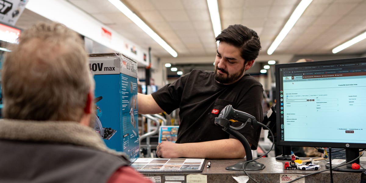 A bearded employee at the customer service counter is helping an elderly customer, holding up what appears to be a blue produ