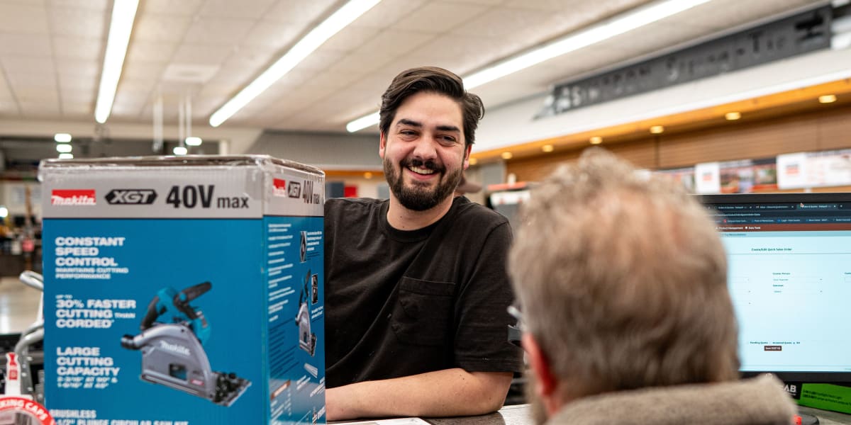 A smiling bearded male employee in a black shirt is helping a customer at what appears to be the customer service counter or